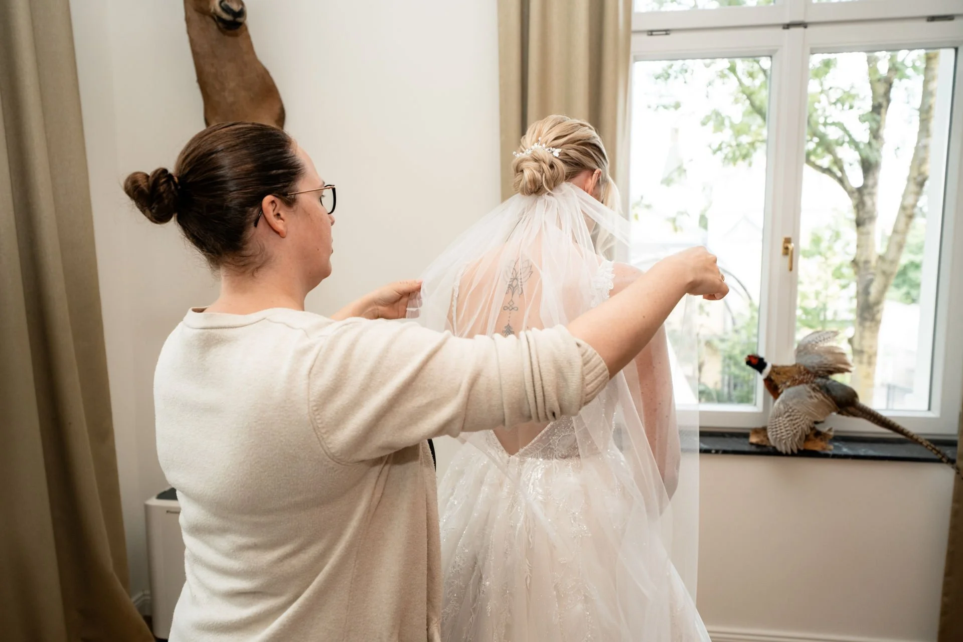 Eine Braut bekommt Hilfe beim Anziehen ihres Hochzeitskleides in einem Schlafzimmer mit großen Fenstern und Naturblick. Eine Frau assistiert, während die Braut einen Schleier trägt.