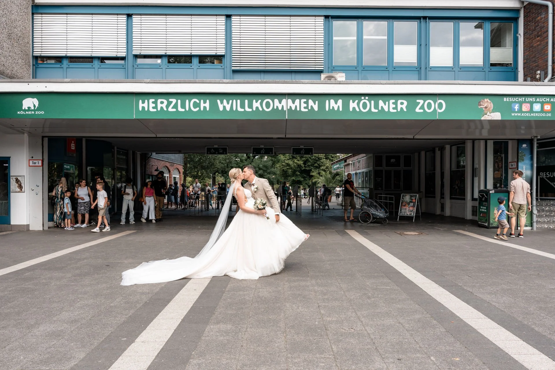 Ein Brautpaar, das sich küsst, im Vordergrund, vor dem Eingang des Kölner Zoos, mit vielen Besuchern im Hintergrund.