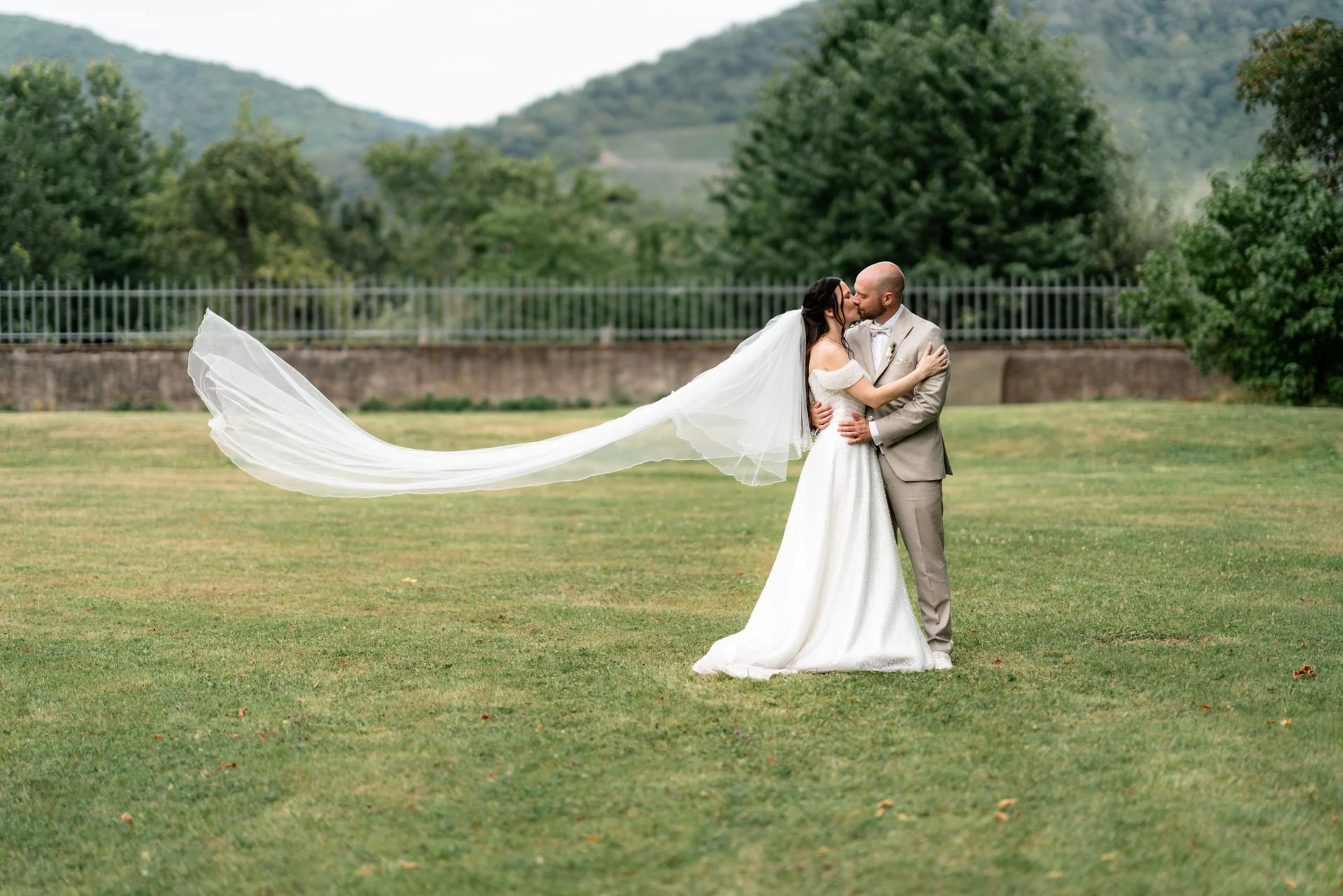Ein Brautpaar küsst sich auf einer Wiese im Freien, die Braut trägt ein weißes Kleid mit Schleier, der sich im Wind bewegt, und der Bräutigam einen hellen Anzug, im Hintergrund sind Bäume und Berge sichtbar.