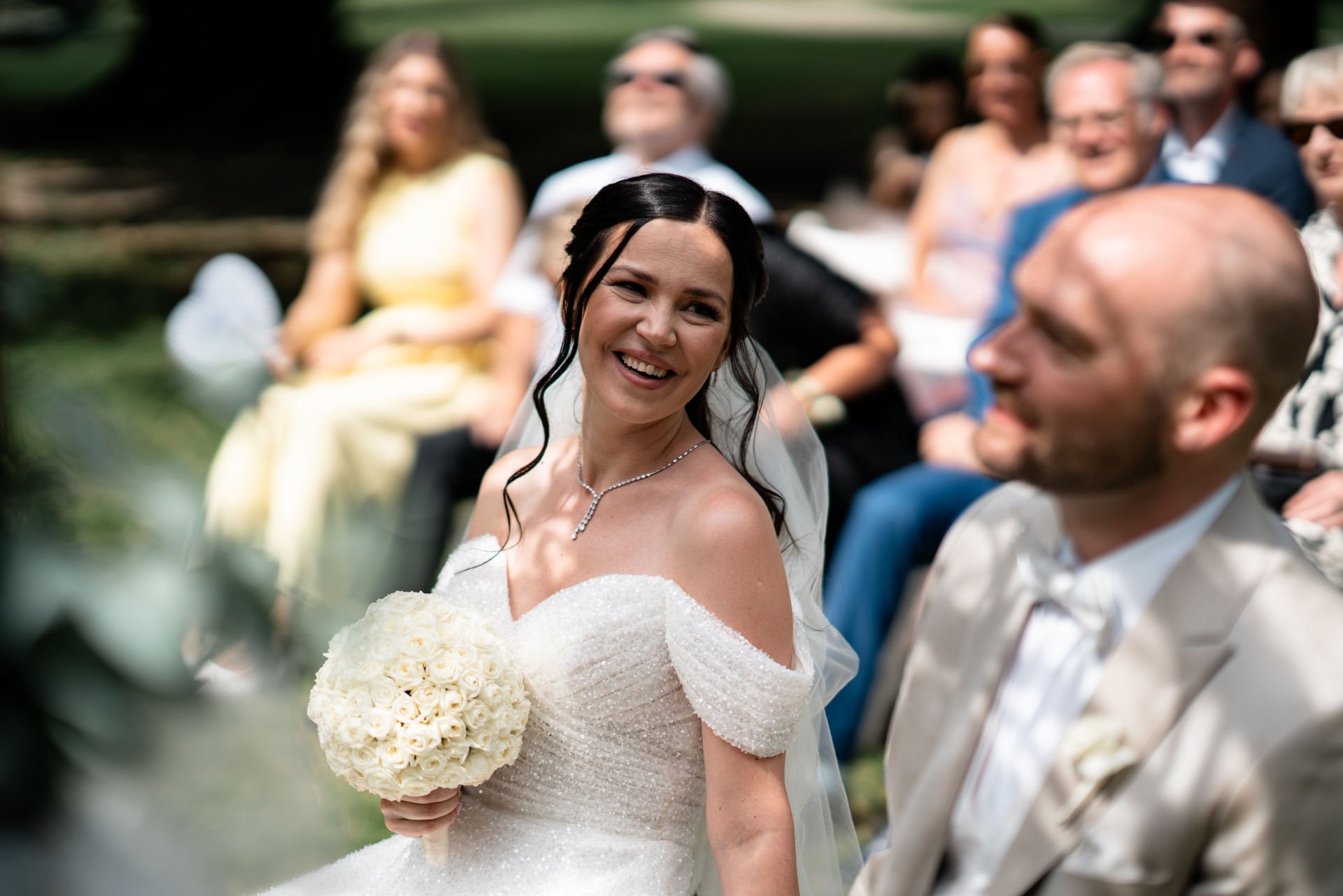 Eine gepflegte Frau in Brautkleid, die auf einer Hochzeit lachend neben einem Mann sitzt, umgeben von Freunden und Familienmitgliedern im Freien.