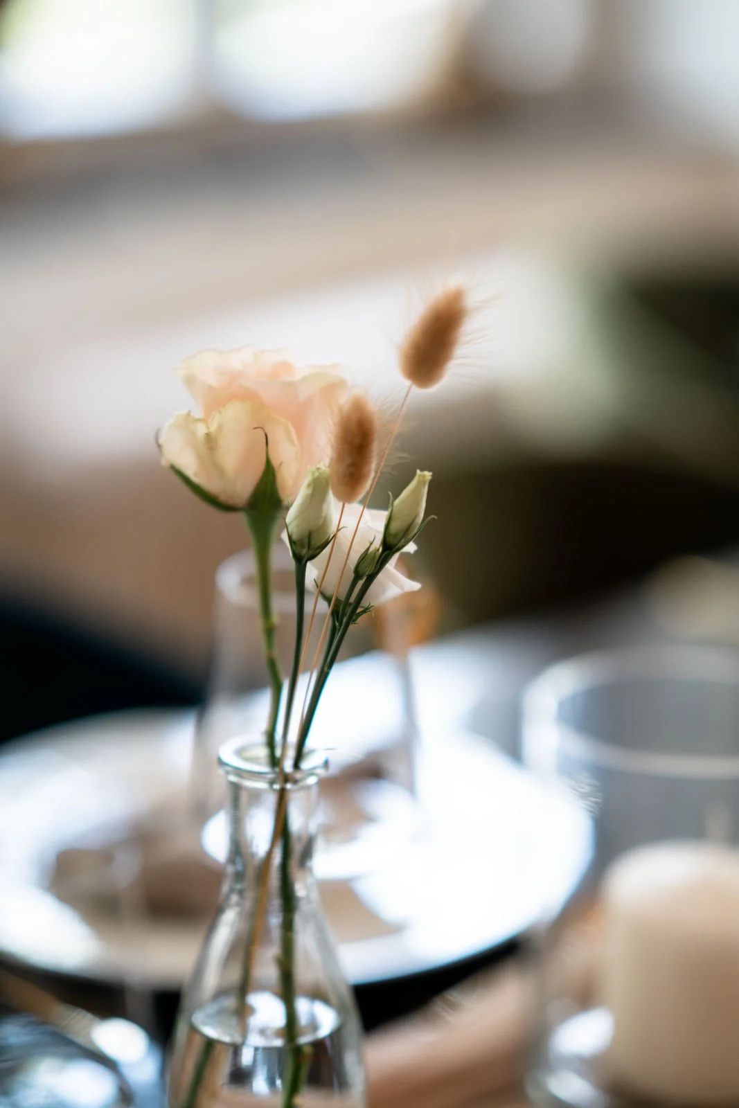 Eine kleine Glasvase mit zarten Blumen und Grashalmen, im Hintergrund unscharf eine Tischsituation.