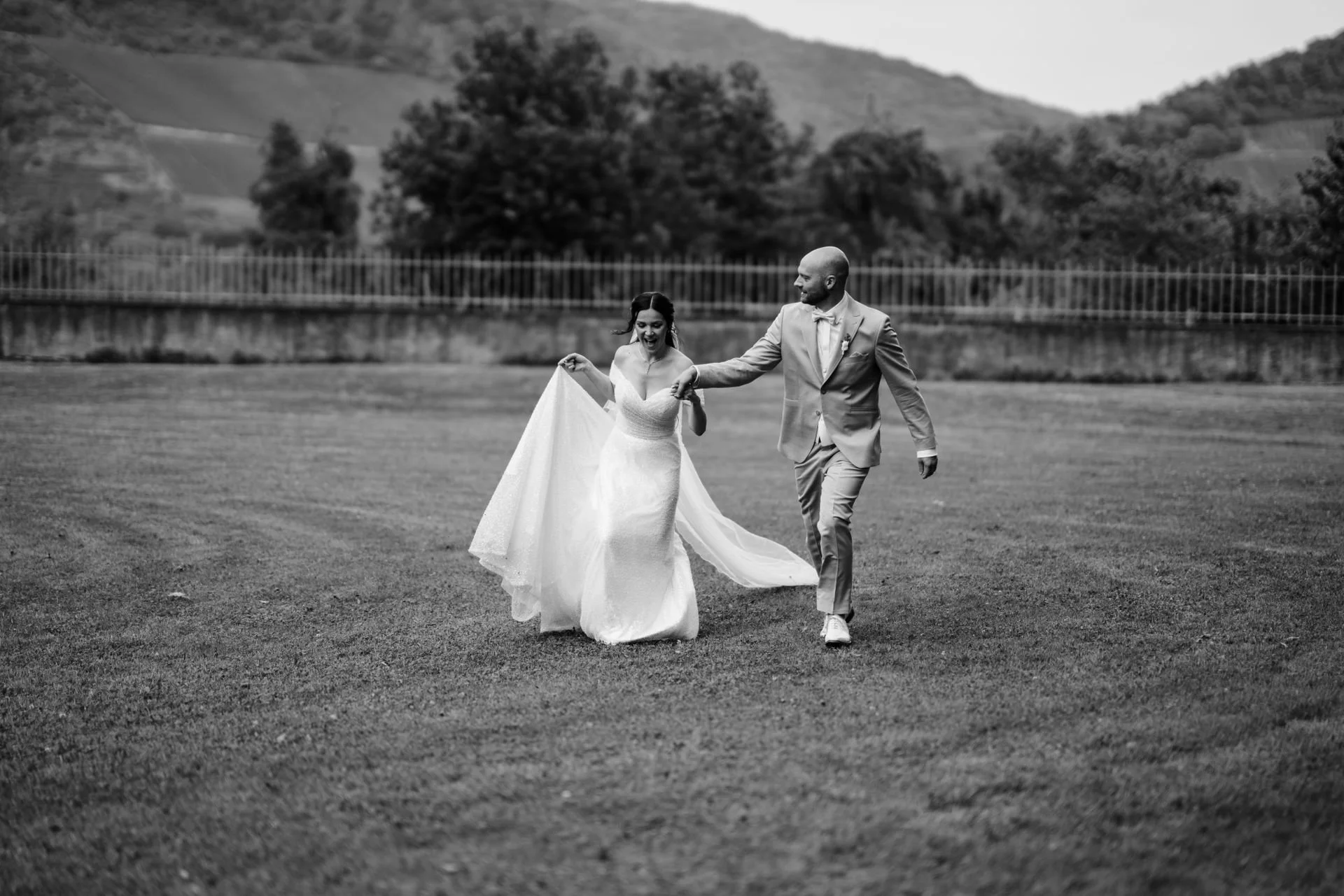 Braut und Bräutigam tanzen und lachen im Freien, umgeben von Hügeln und Bäumen, in einem schwarzen und weißen Hochzeitsbild.