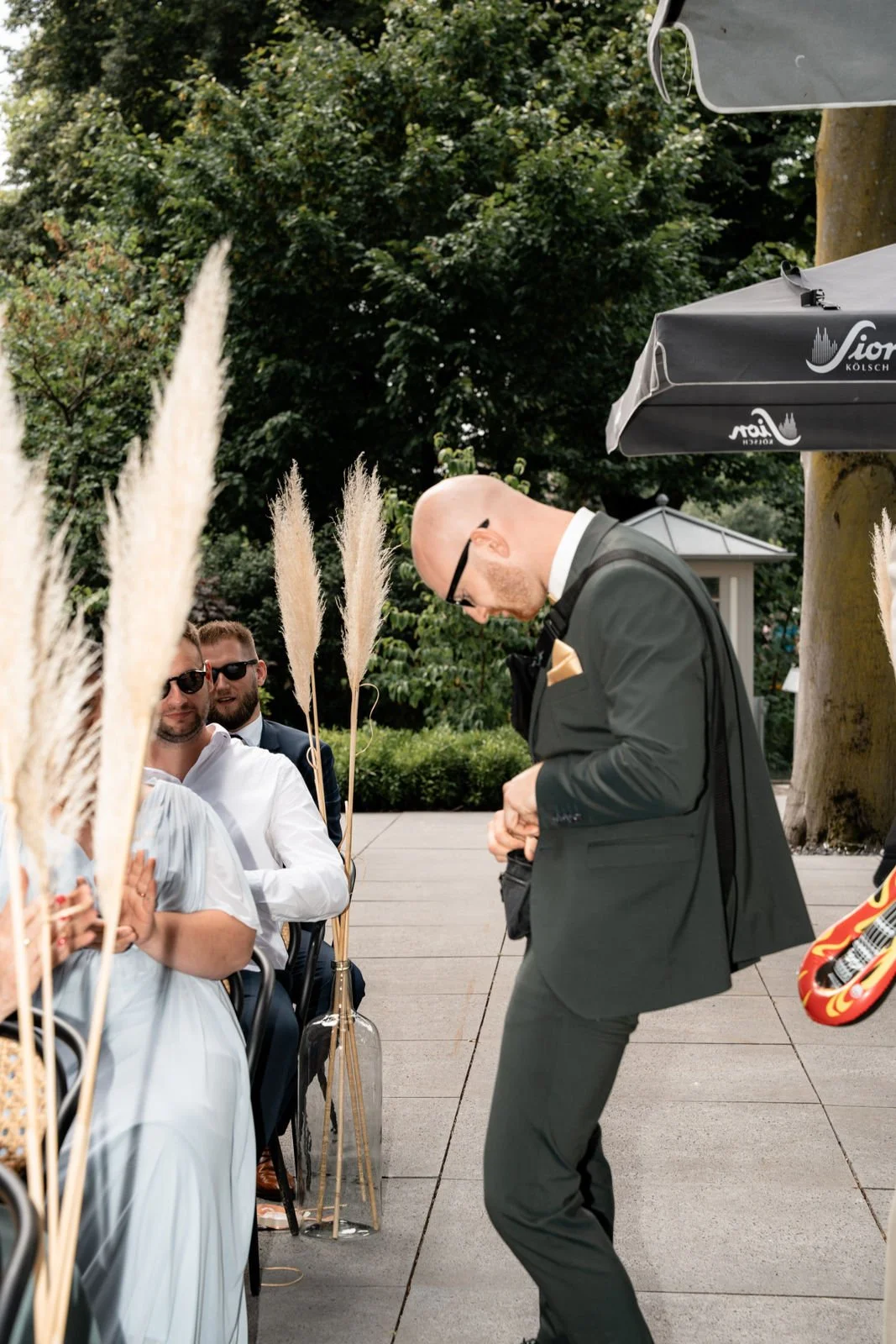 Mann in smoking mit Brille, der vor einer Gruppe sitzt, während eine Person im grauen Anzug und mit Brille auf dem Boden steht und sich die Taschen richtet. Zwei andere Männer sitzen im Hintergrund, einer trägt Sonnenbrille. Es ist ein Garten oder ei
