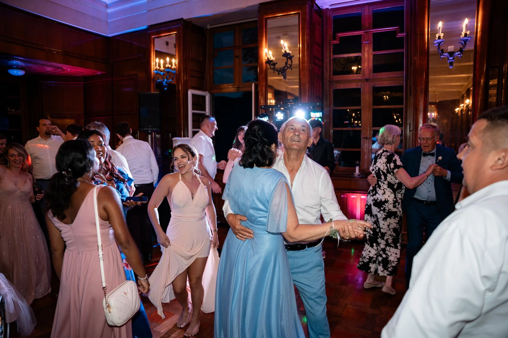 Menschen tanzen und feiern in einem festlichen Raum mit Holzvertäfelung und Kronleuchtern.