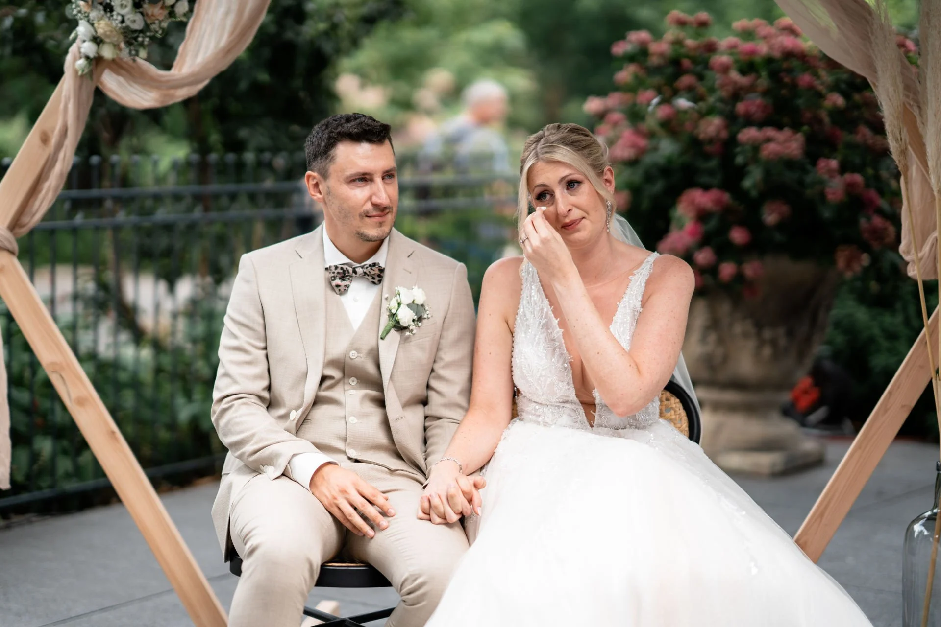 Ein Hochzeitspaar sitzt bei einer emotionalen Zeremonie im Freien, der Bräutigam hält die Hand der weinenden Braut. Im Hintergrund blühen Pflanzen und Blumen.