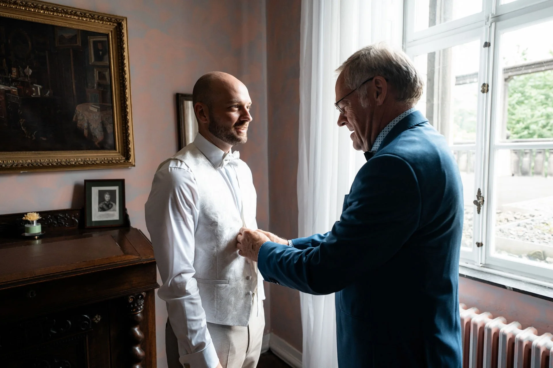 Ein jüngerer Mann in weißer Trauungskleidung erhält von einem älteren Mann in dunklem Anzug eine Anstecknadel, wahrscheinlich für die Hochzeit, in einem Raum mit einem großen Fenster, einem Gemälde und einem kleinen Porträt auf einem Schreibtisch.