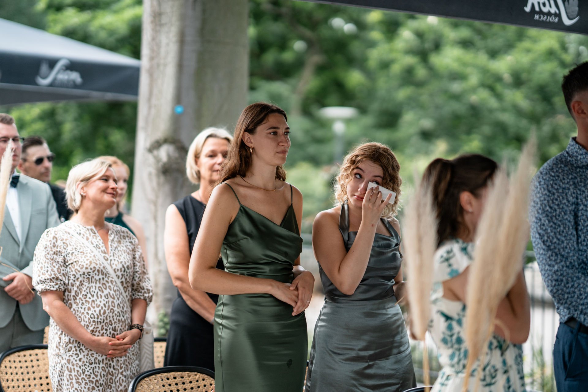Menschen bei einer formellen Veranstaltung im Freien, einige weinen, andere trösten sie, umgeben von grüner Natur.