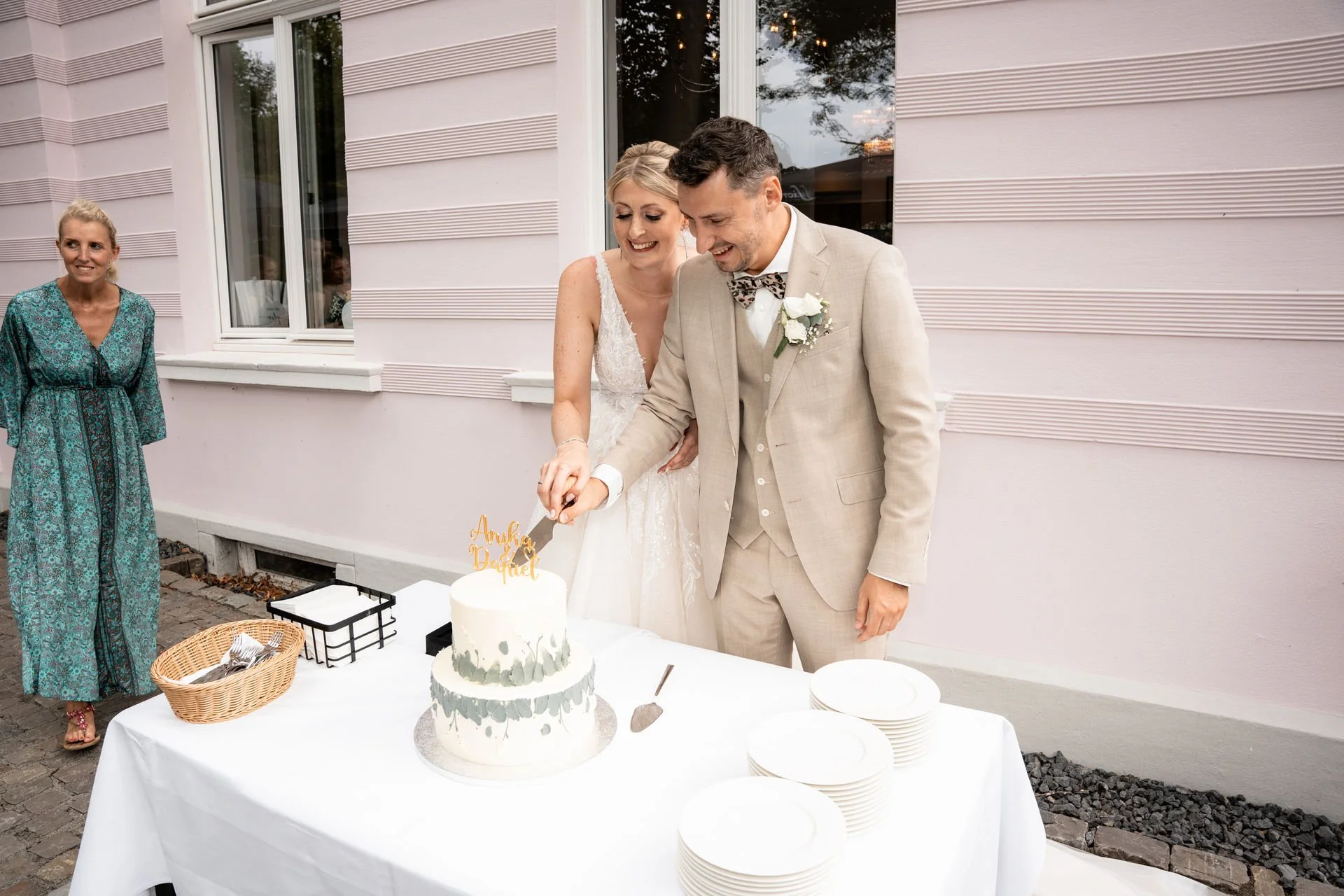 Ein Brautpaar schneidet auf einer Hochzeitstorte zu. Die Mutter der Braut steht im Hintergrund und beobachtet. Die Torte ist weiß mit grauen Wasserfarben-Designs und einem goldenen Hochzeitsdekor. Es liegen Teller und Besteck bereit.
