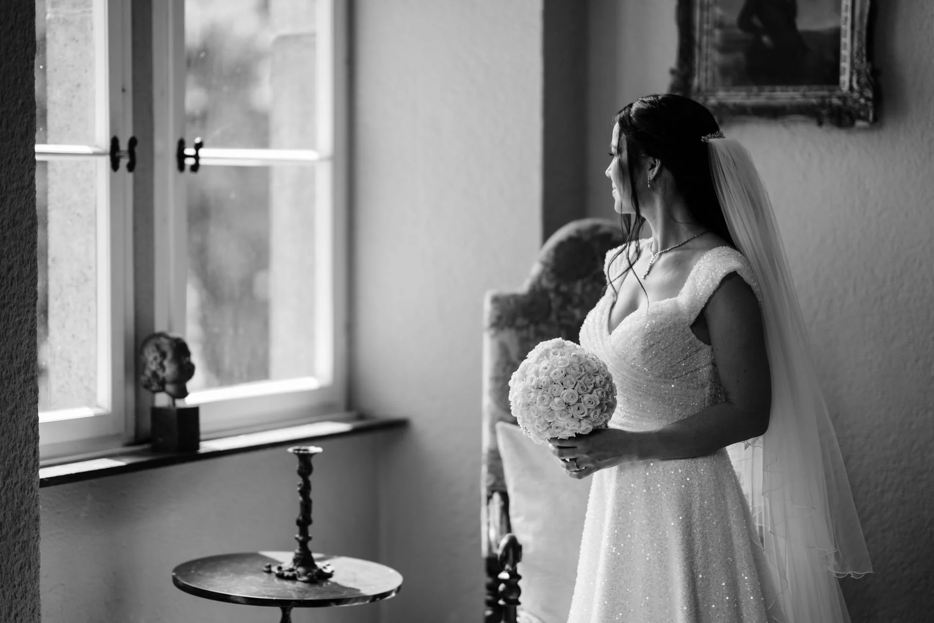 Eine Braut in einem weißen, funkelnden Brautkleid hält einen Bouquet aus Rosen und steht neben einem Fenster, während sie nach draußen schaut.