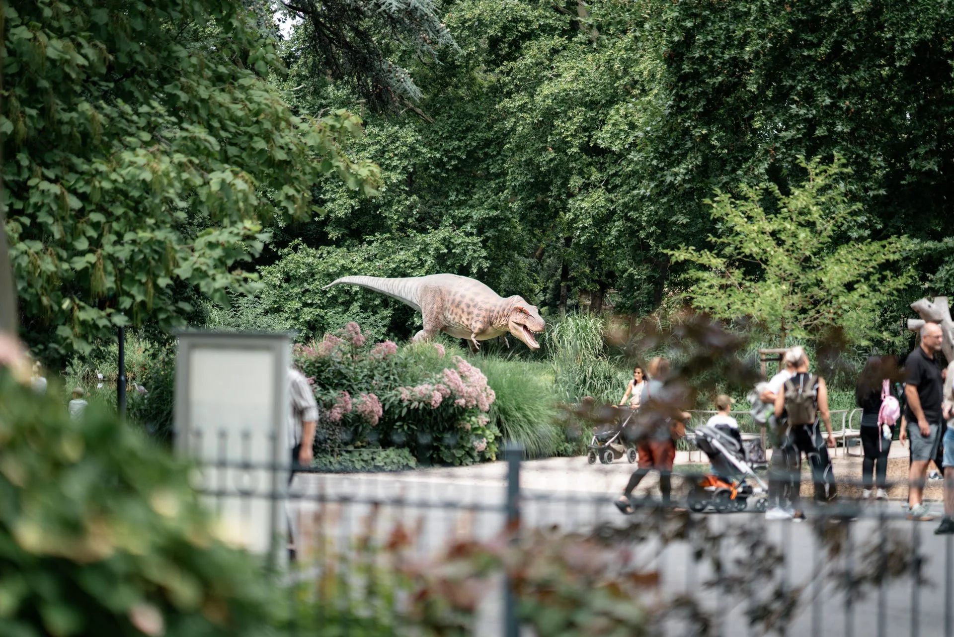 Menschen in einem Zoo, unter anderem ein Mädchen mit Kinderwagen, während ein Dinosaurier-Attrappe im Hintergrund sichtbar ist.