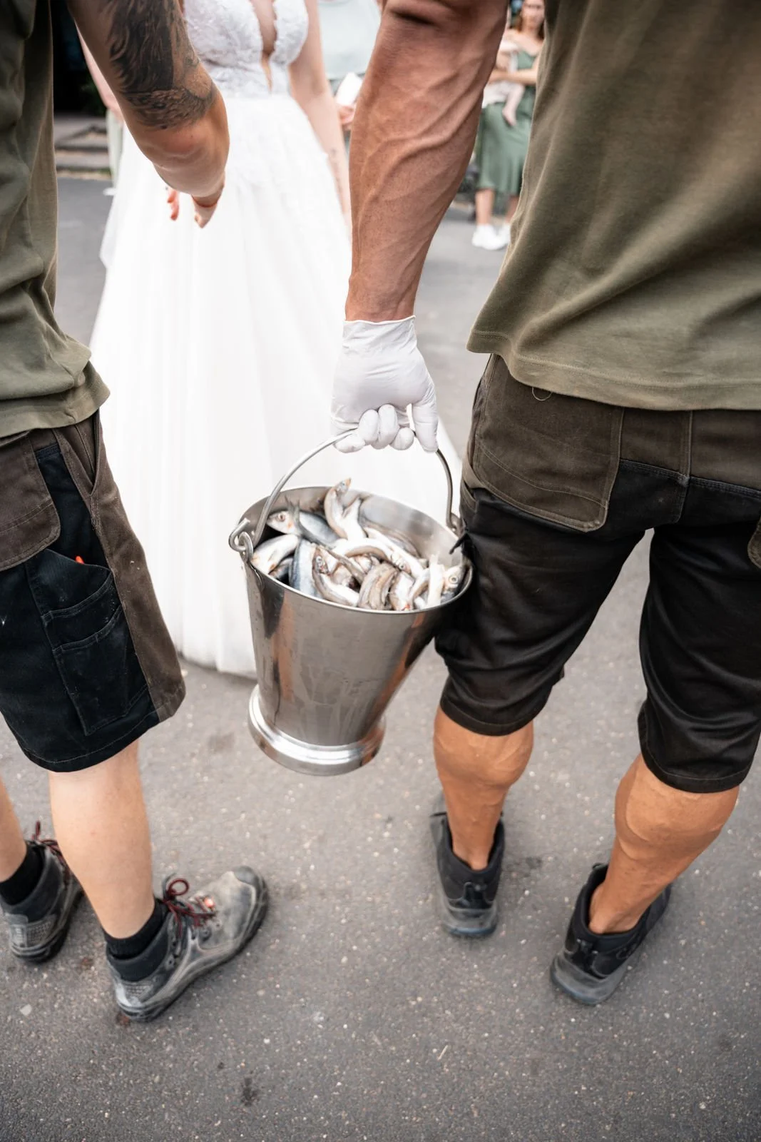 Zwei Männer stehen im Freien, einer hält einen Korb mit frisch gefangenen kleinen Fischen. Im Hintergrund sind Menschen, darunter eine Frau in einem weißen Kleid, sichtbar.