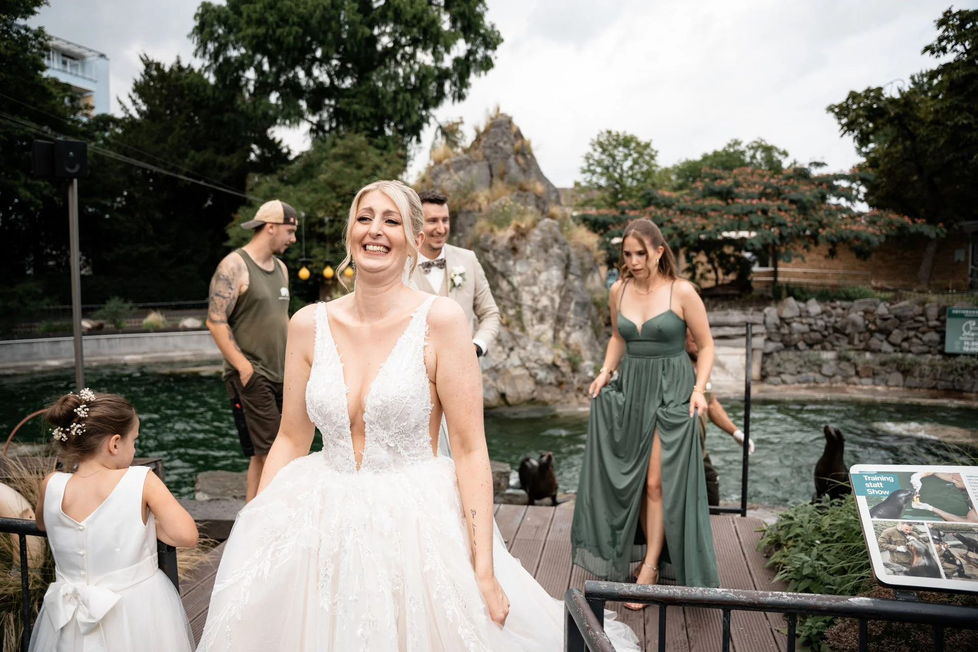 Eine glückliche Braut in einem weißen Brautkleid mit mehreren Gästen im Hintergrund bei einer Hochzeit im Freien am Wasser mit Felsen und Bäumen.