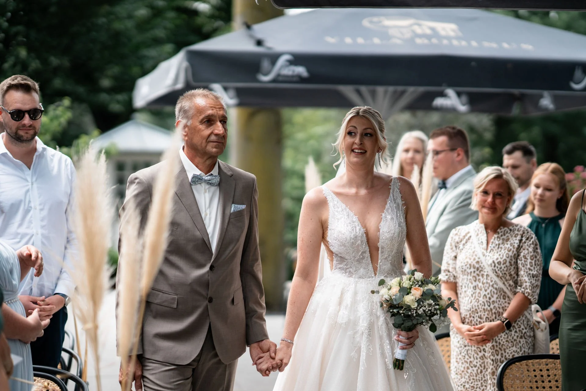 Braut in einem weißen Hochzeitskleid hält die Hand eines Mannes in einem grauen Anzug bei einer Eheschließung im Freien.