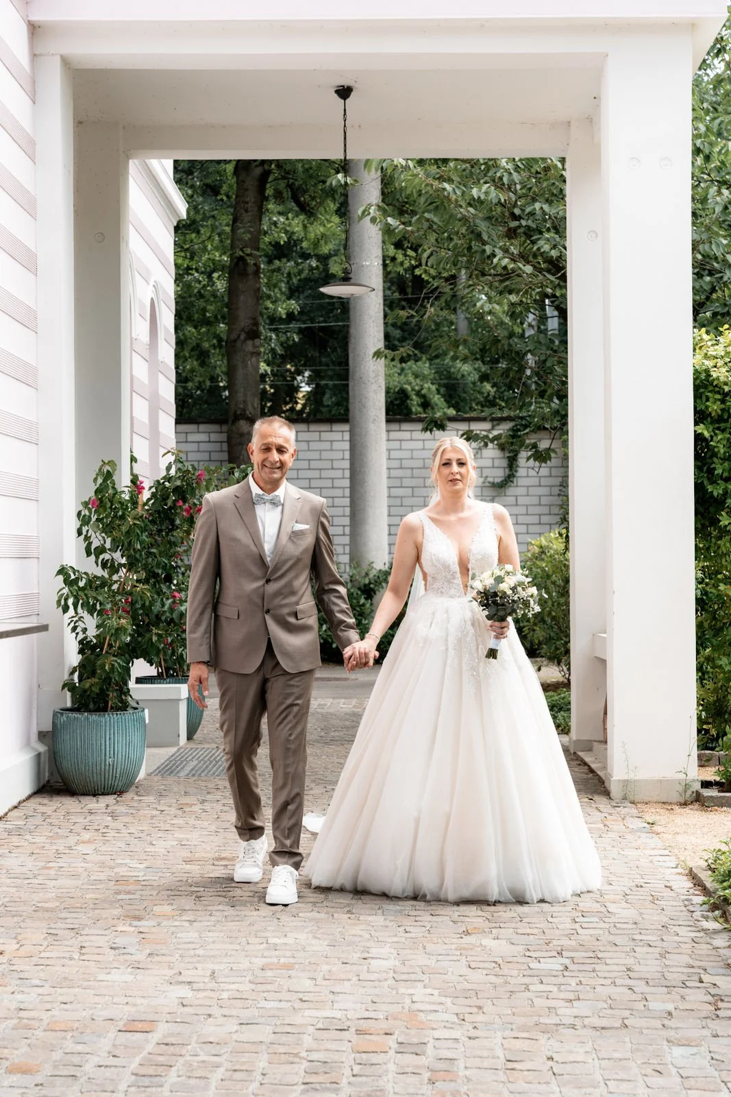 Braut in Hochzeitskleid und Bräutigam in Anzug halten Händchen und gehen auf einem gepflasterten Weg. Im Hintergrund sind grüne Bäume, eine weiße Mauer und Pflanzen in Töpfen.