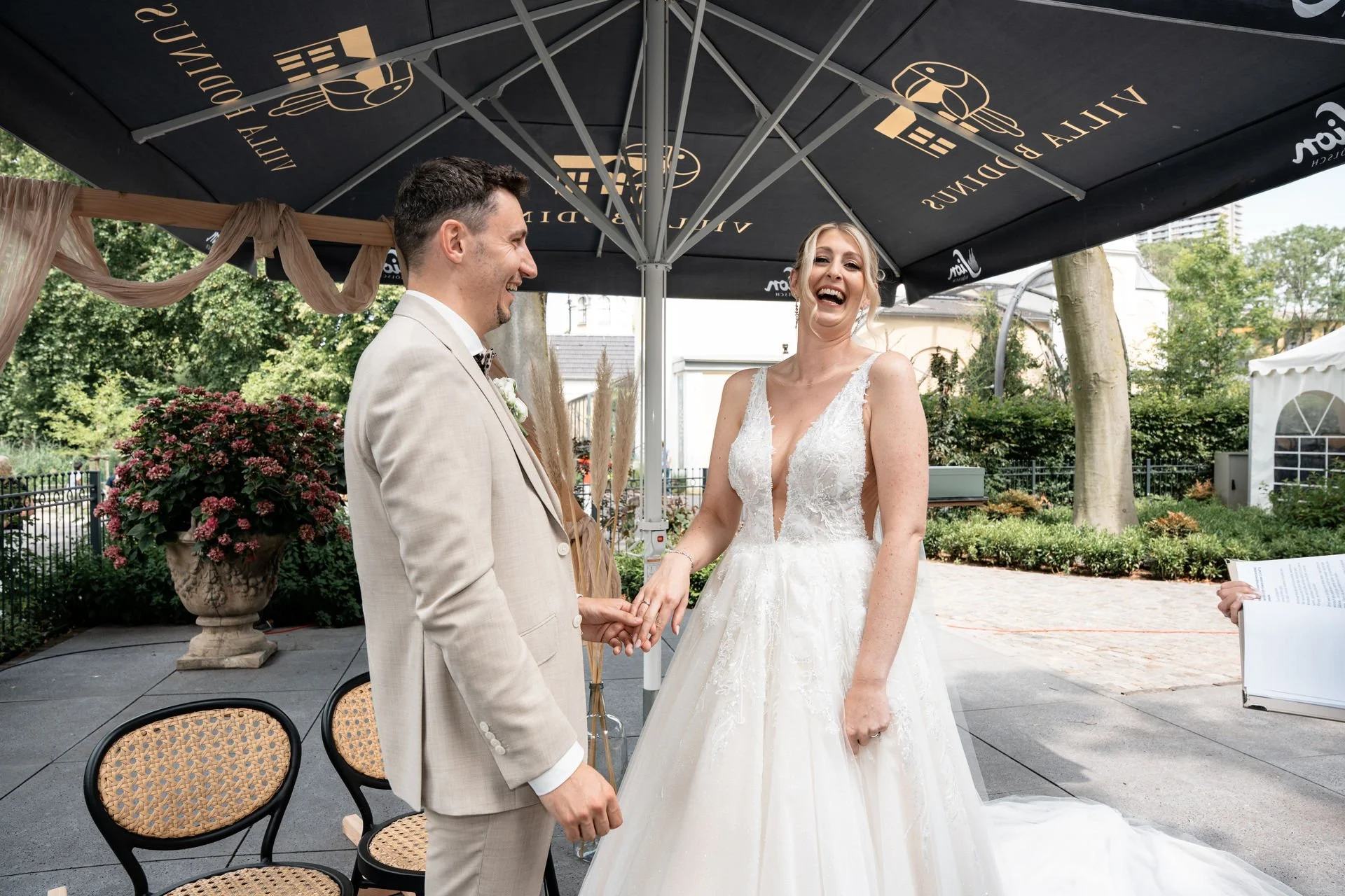 Ein glückliches Brautpaar während ihrer Hochzeit im Freien, unter einem großen schwarzen Sonnenschirm, umgeben von Grünpflanzen und Sitzgelegenheiten, beim Austausch von Eheringen.