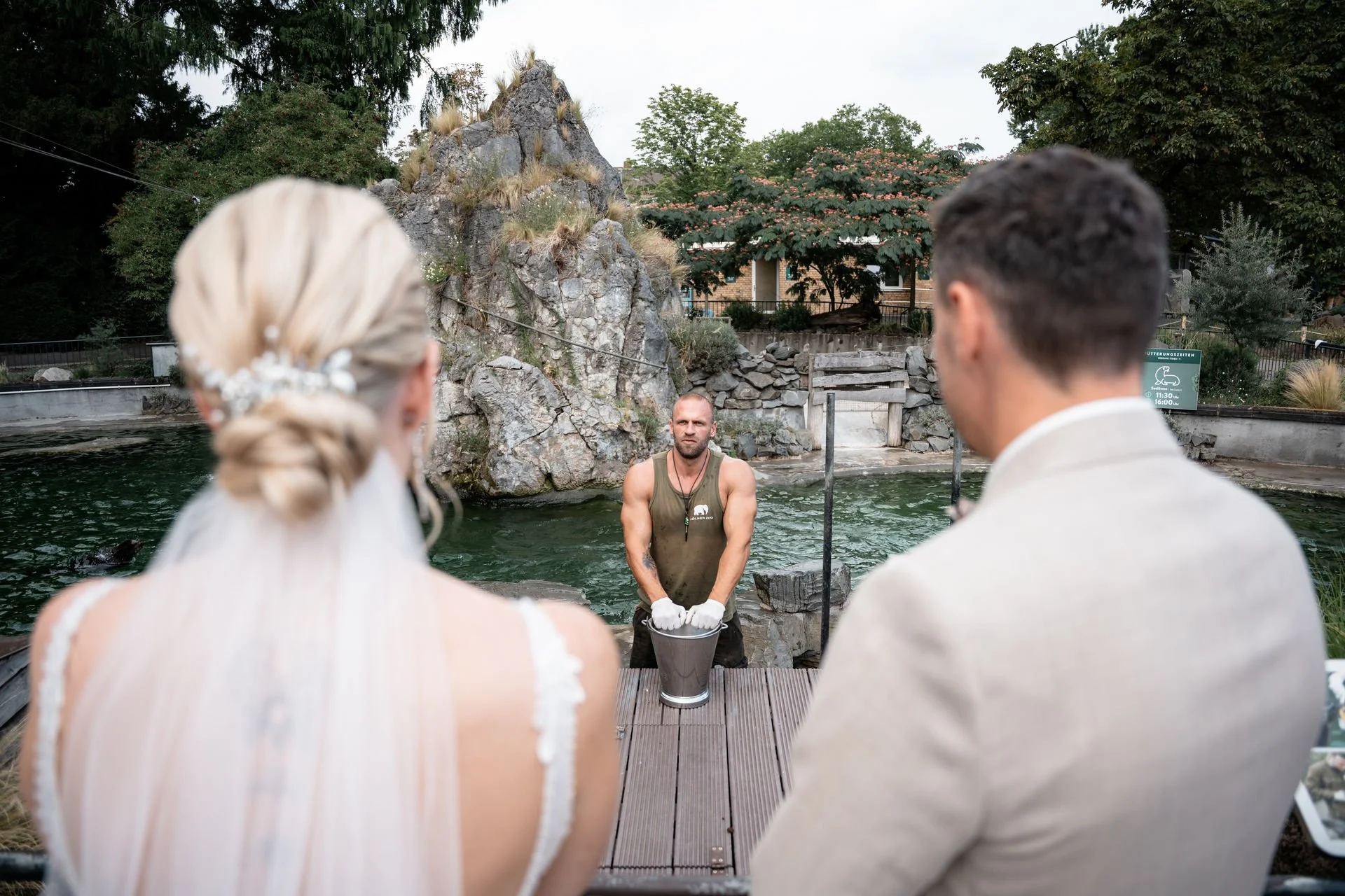 Ein Brautpaar bei ihrer Trauung im Freien am Wasser, der Zeremonienleiter steht in der Mitte mit traurigem Gesicht und hält eine Schüssel. Der Hintergrund zeigt einen Wasserfall und Bäume.
