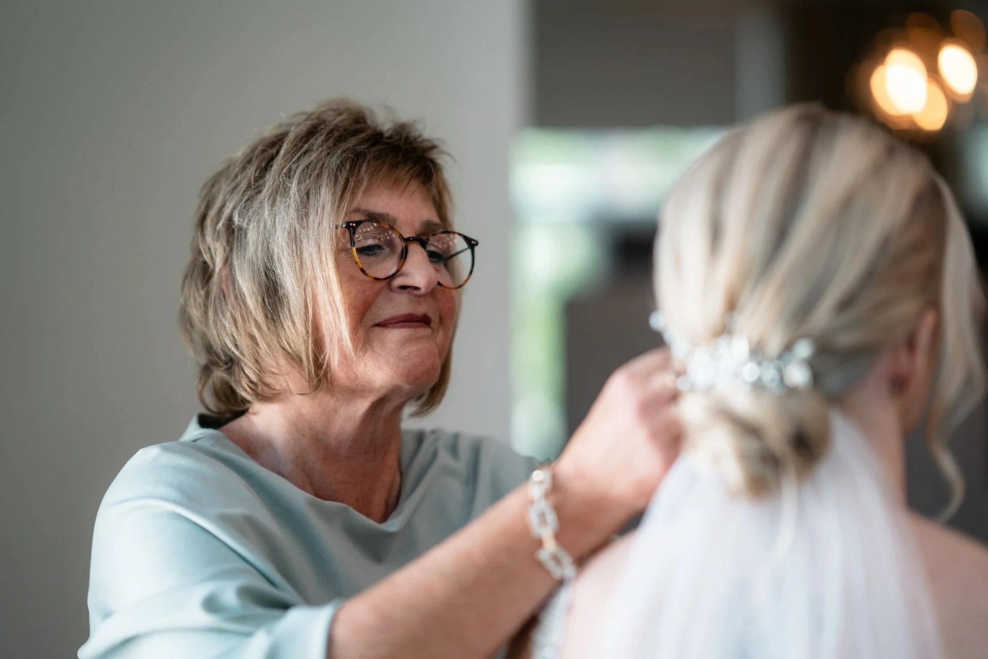 Eine ältere Frau mit Brille richtet die Haare einer jüngeren Frau für eine Hochzeit oder Feier. Im Hintergrund sind warme Lichtquellen sichtbar.