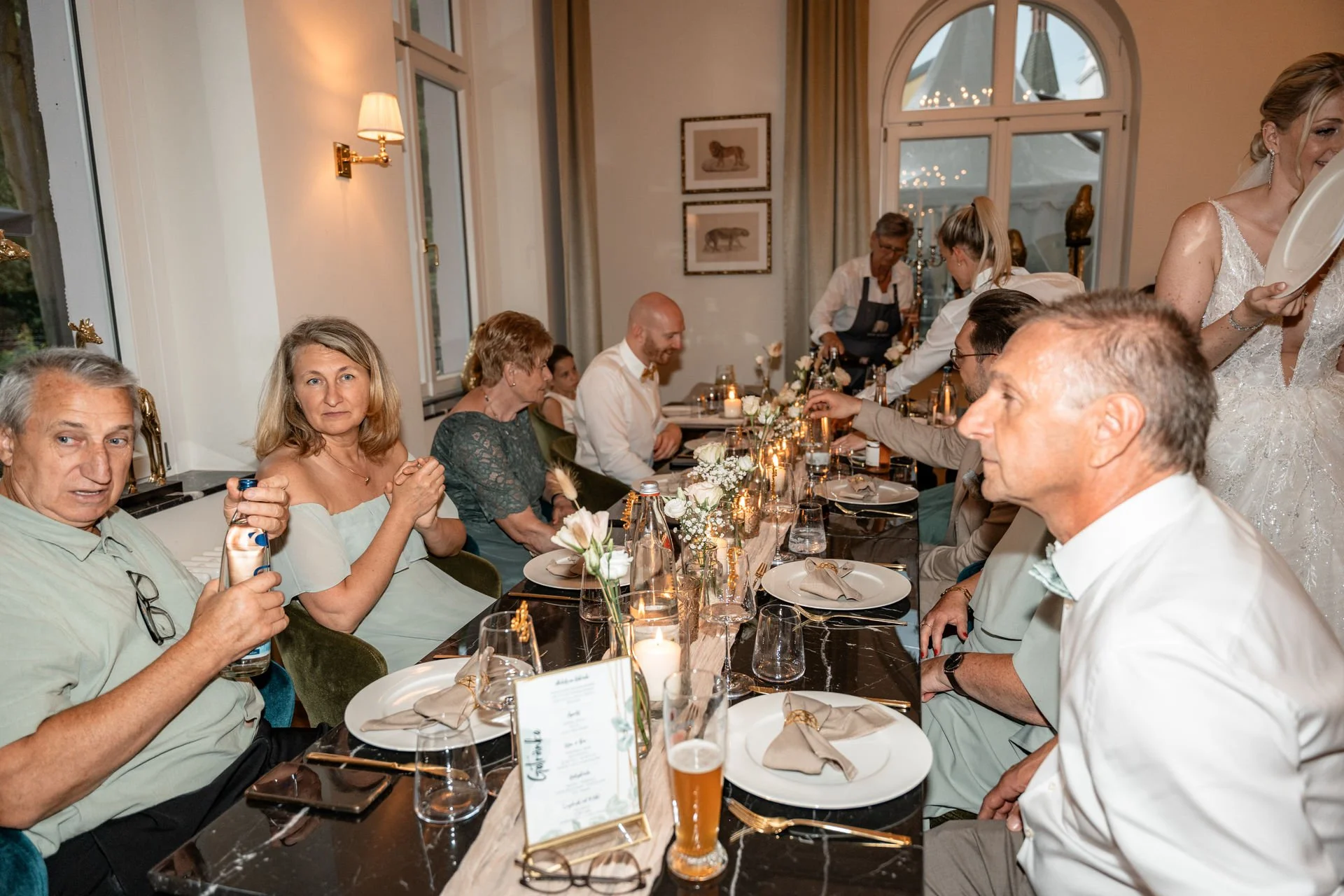Menschen sitzen an einem festlich gedeckten Tisch bei einer Feier, wahrscheinlich einer Hochzeit, in einem hell erleuchteten Raum mit Fenstern und Wanddekorationen, einige Personen trinken Getränke, andere unterhalten sich.