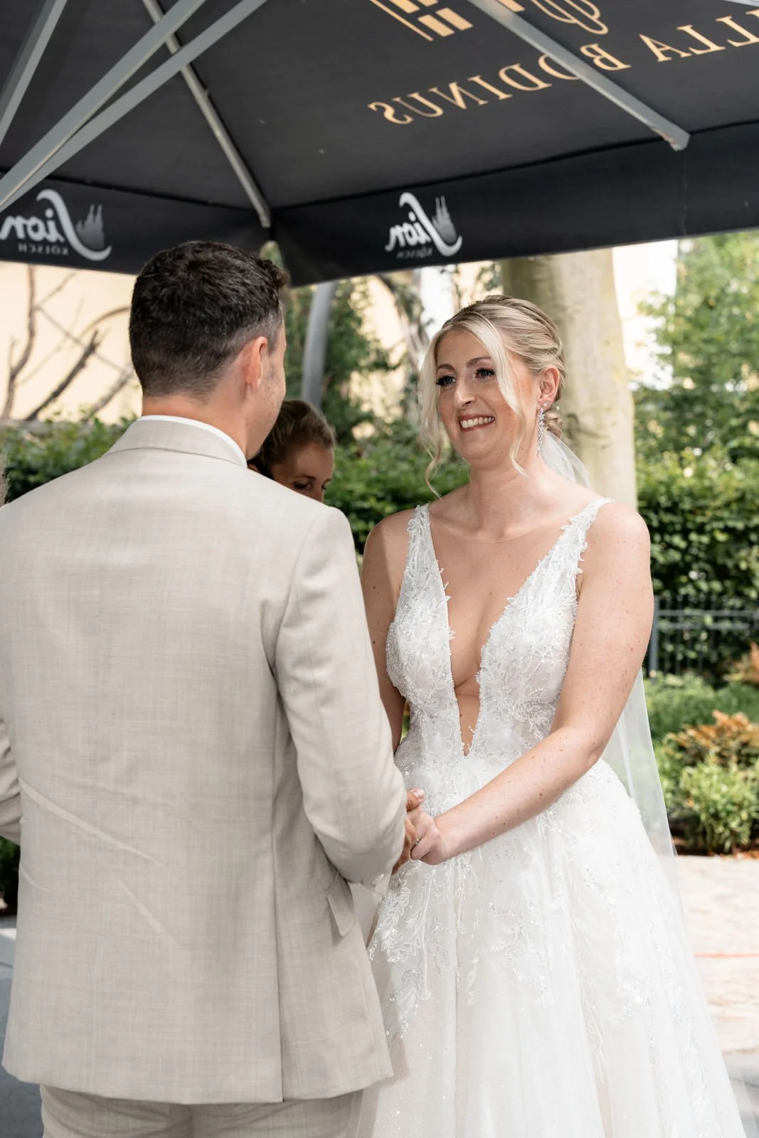 Braut und Bräutigam bei Hochzeitszeremonie im Freien, die Braut trägt ein weißes Kleid mit Spaghettiträgern, während der Bräutigam einen hellen Anzug trägt, unter einem schwarz-weißen Sonnenschirm.
