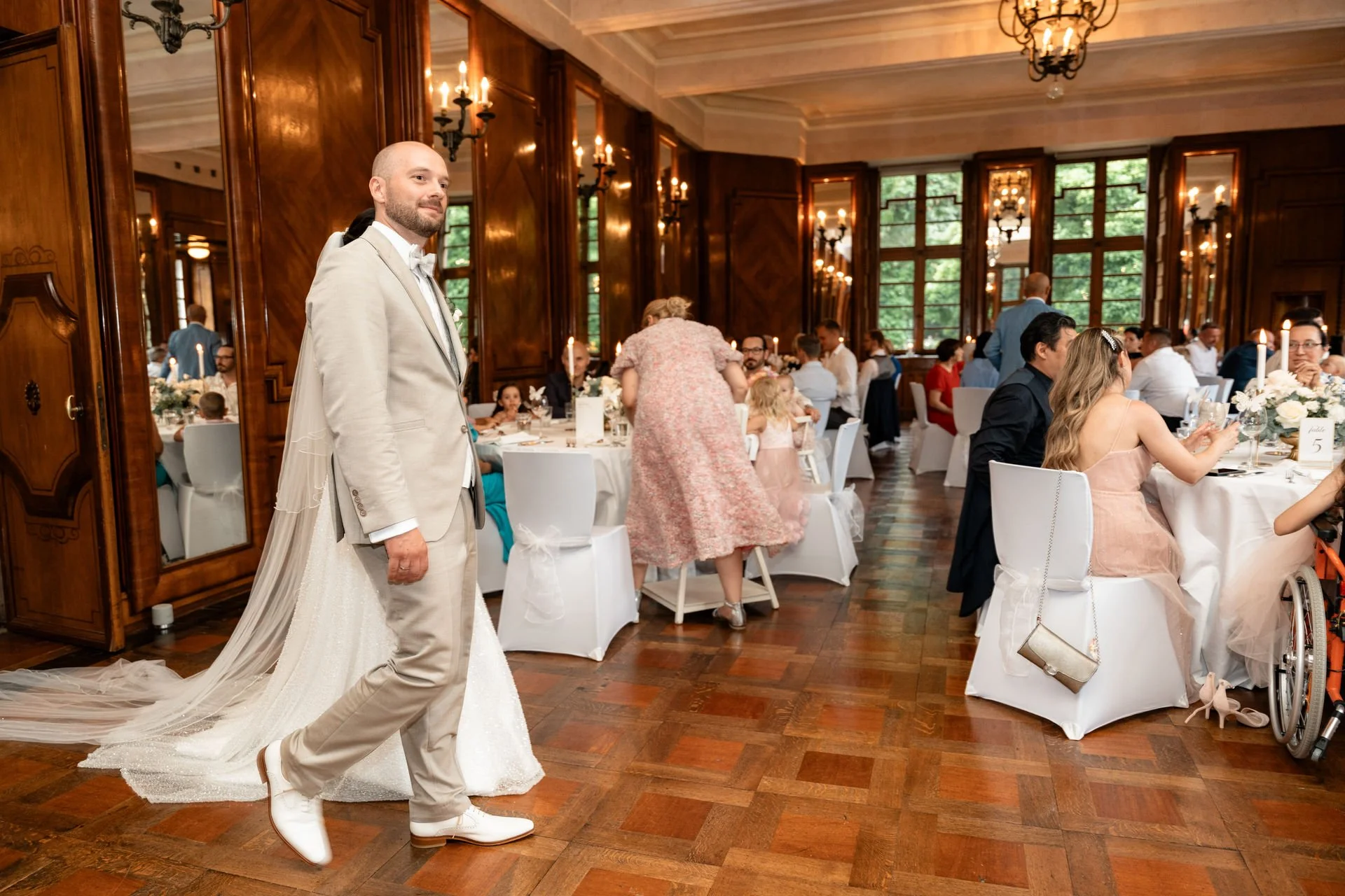Ein Bräutigam in einem cremefarbenen Anzug möchte in einem festlich geschmückten Saal auf einer Hochzeit sitzen. Im Hintergrund sind Gäste an Tischen mit weißen Tischdecken und Blumen-Dekorationen. Der Raum hat große Fenster, hölzerne Wände und Kronl