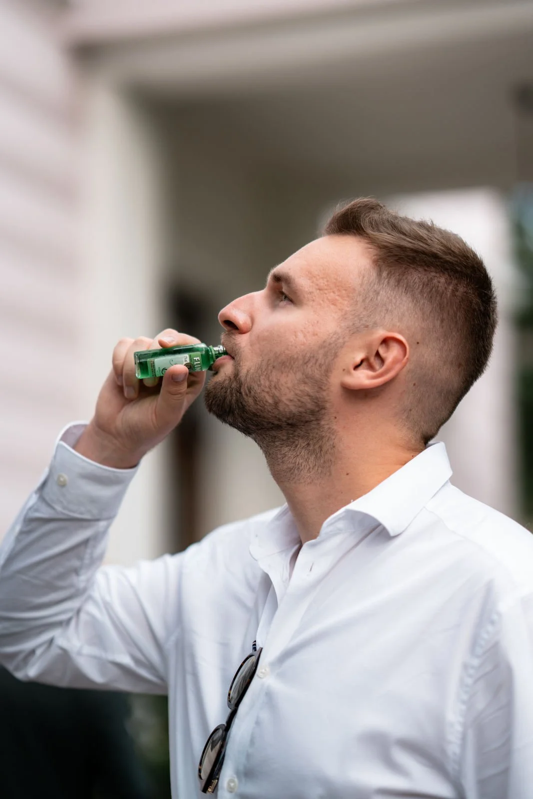Ein Mann in Weißes Hemd trinkt aus einer kleinen grünen Flasche auf einer Terrasse im Freien.