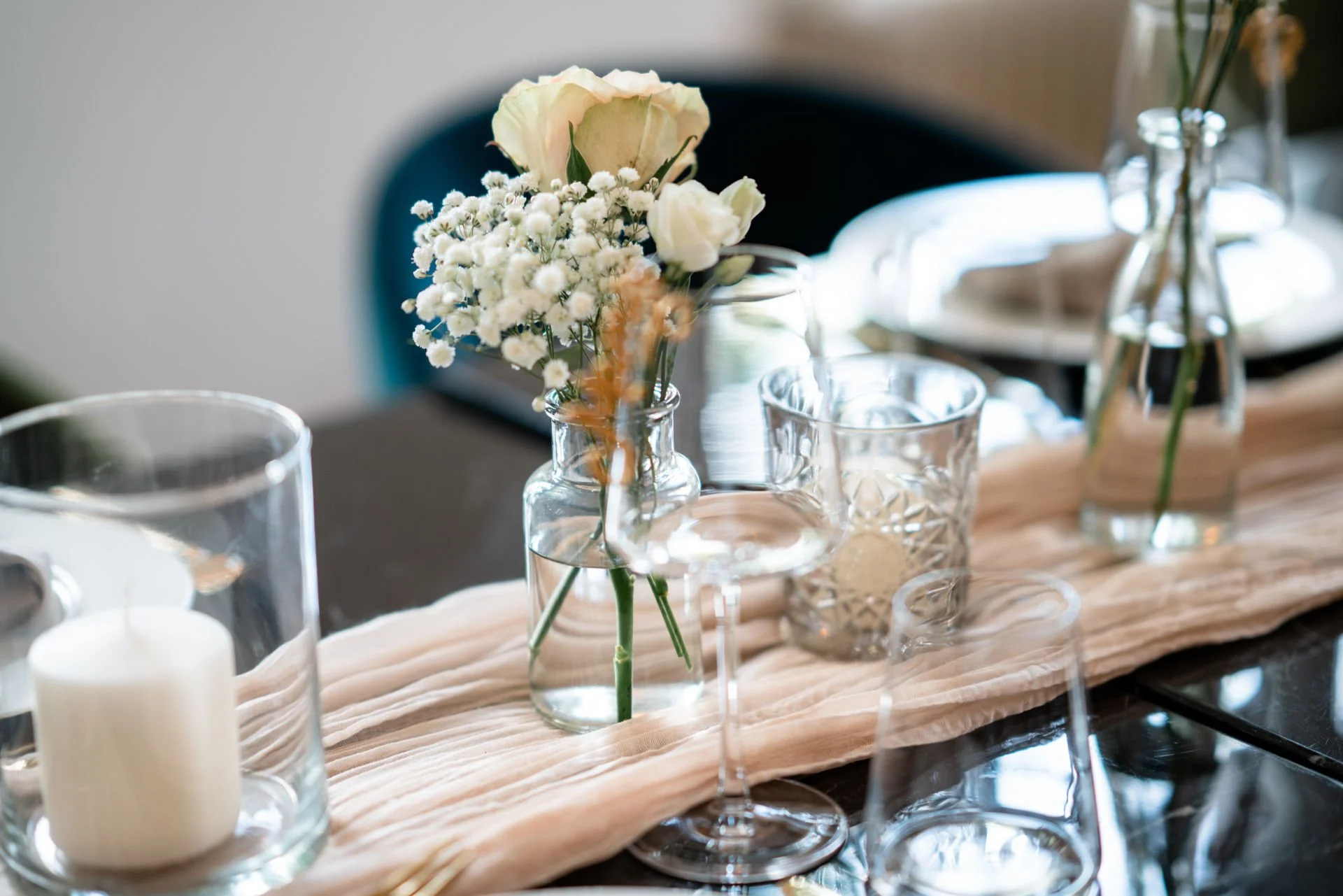 Blumenarrangement mit weißen Rosen, Baby's Breath und grünen Blättern in einer Glasvase auf einem Tisch, umgeben von Gläsern und Kerzen in Glasvasen