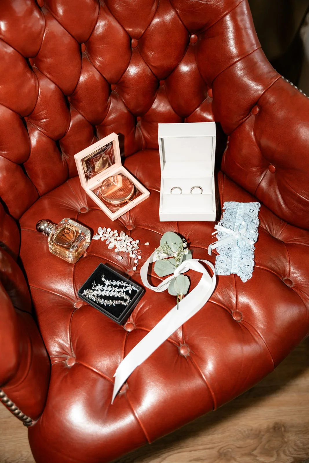 Rotes Ledersofa mit verschiedenen Schmuckstücken, einschließlich Perlenohrringen, einer Tiara, einem Parfümfläschchen, einem Flakon und einer kleinen Trageschleife, die für eine Hochzeit oder ein besonderes Ereignis vorbereitet sind.