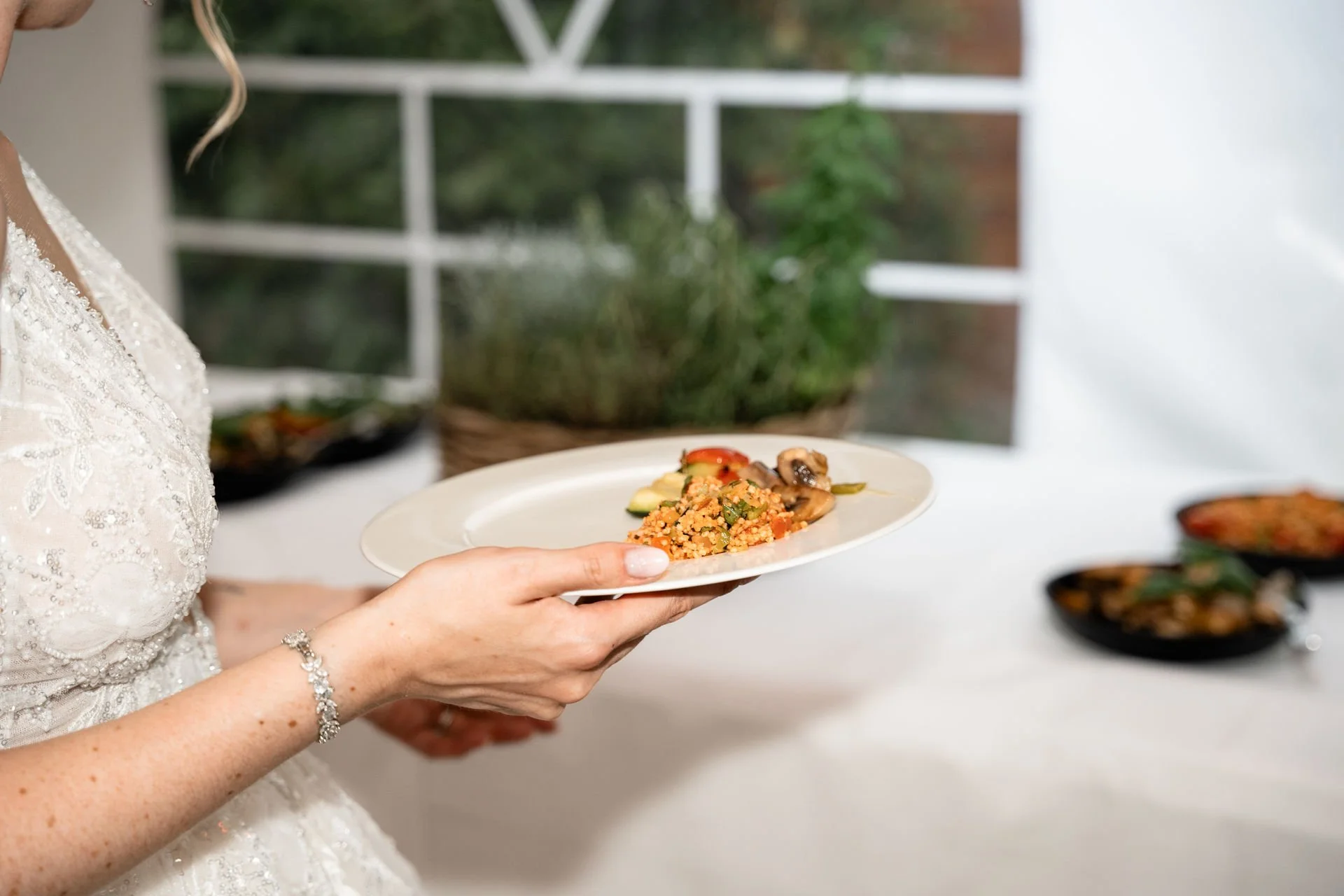 Frau in Hochzeitskleid hält einen weißen Teller mit bunt gemischtem Essen, im Hintergrund Tisch mit weiteren Tellern.