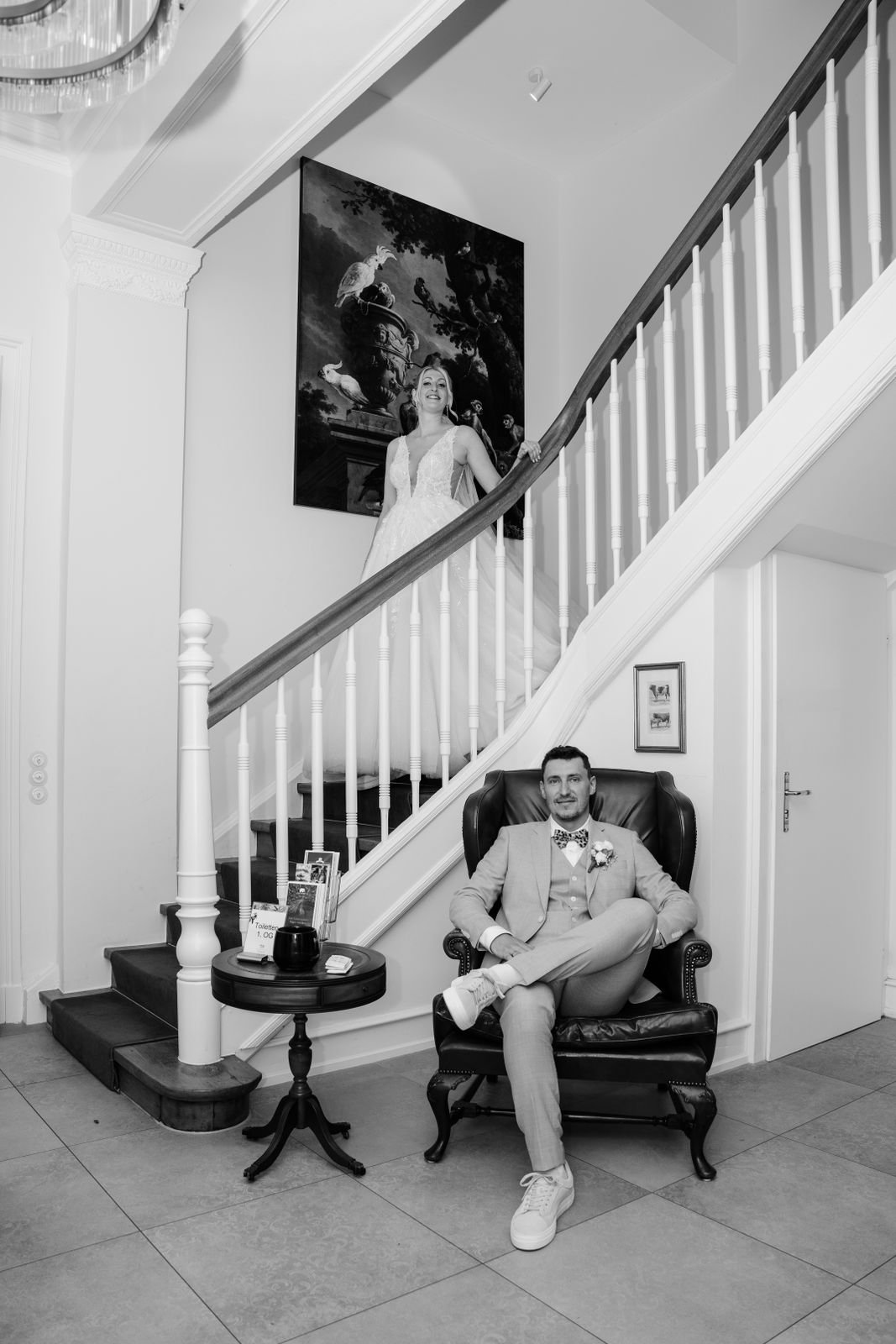 Ein Mann in formeller Kleidung sitzt entspannt in einem schwarzen Ledersessel an einem kleinen Tisch, während eine Braut in einem Hochzeitskleid die Treppe hinaufsteigt.
