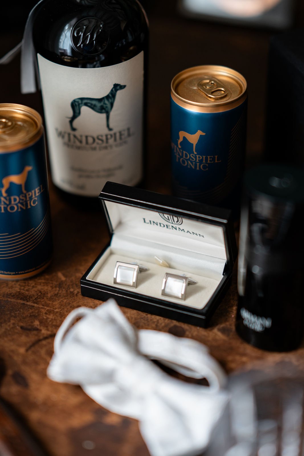 Silberne Manschettenknöpfe in einer schwarzen Schmuckschatulle auf Holztisch, umgeben von Bierdosen, Weinflasche, Gesichtsmaske und einem dunklen Getränkeglas.