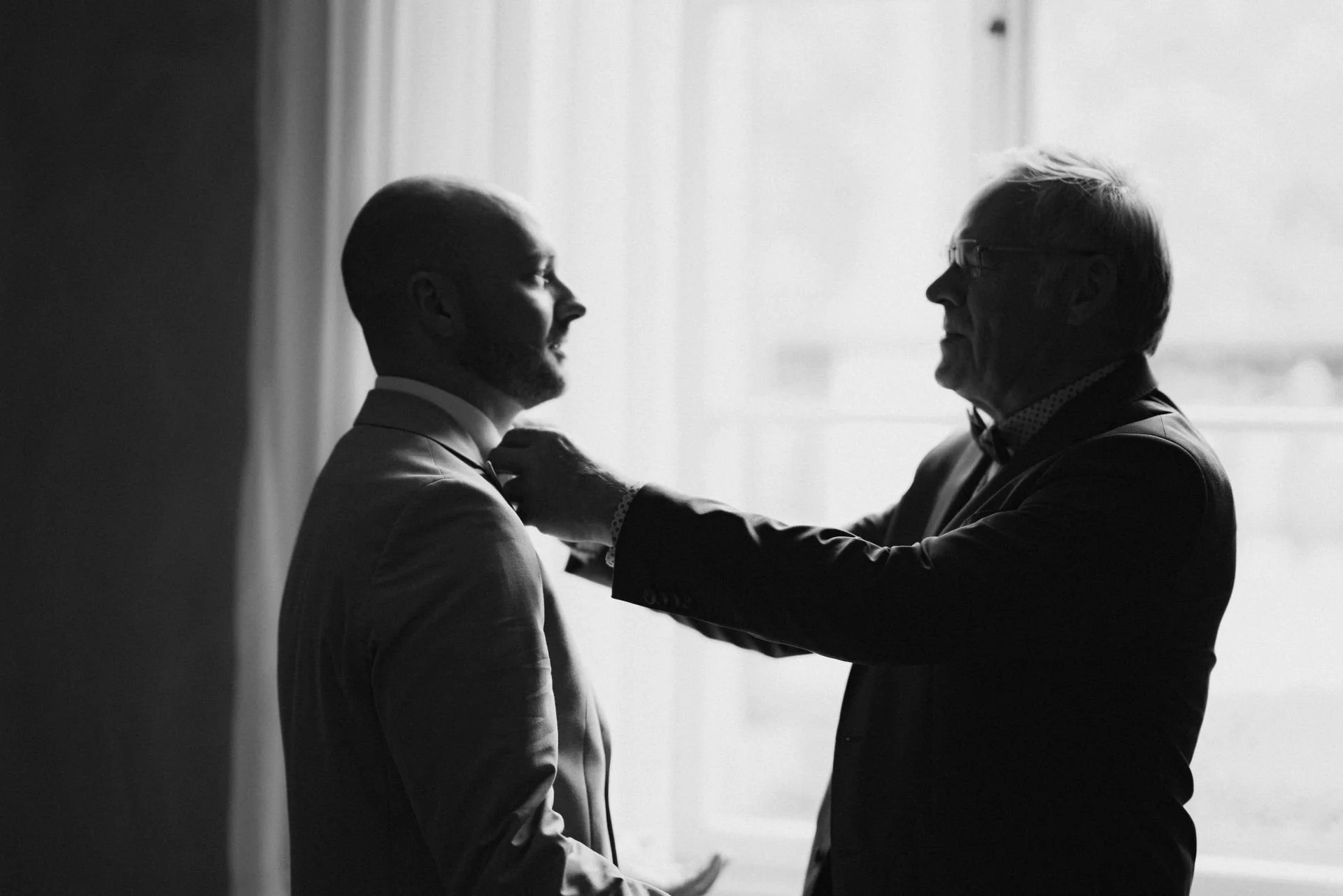 Zwei Männer, einer jüngeren und einer älteren, stehen sich gegenüber im Schatten, beim Anziehen eines Anzugs. Der ältere Mann hilft dem jüngeren, den Kragen richtig zu richten.