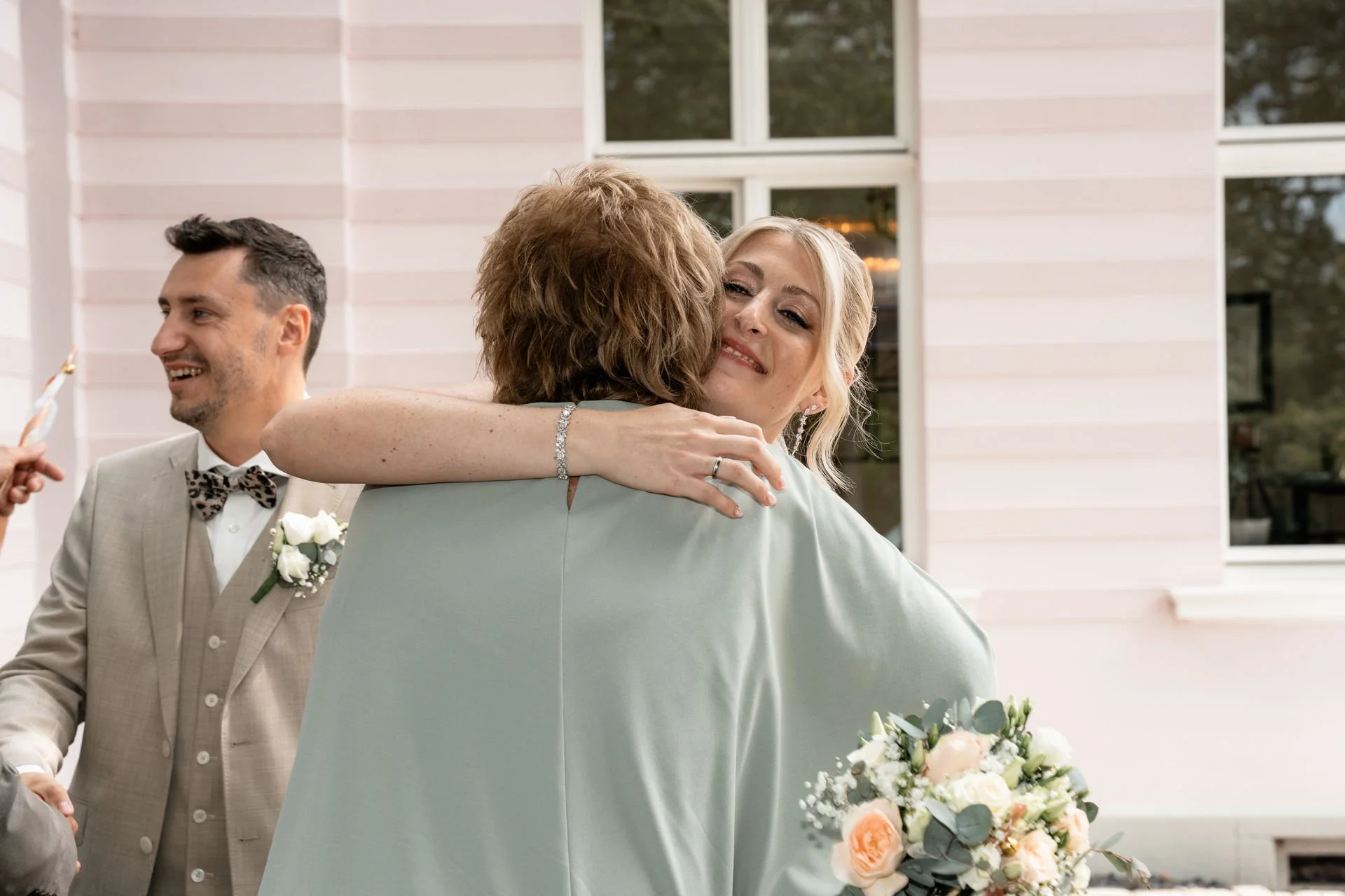 Frau umarmt andere Person bei einer Feier, während ein Mann im Hintergrund lächelnd zuschaut, hält einen Blumenstrauß in der Hand.