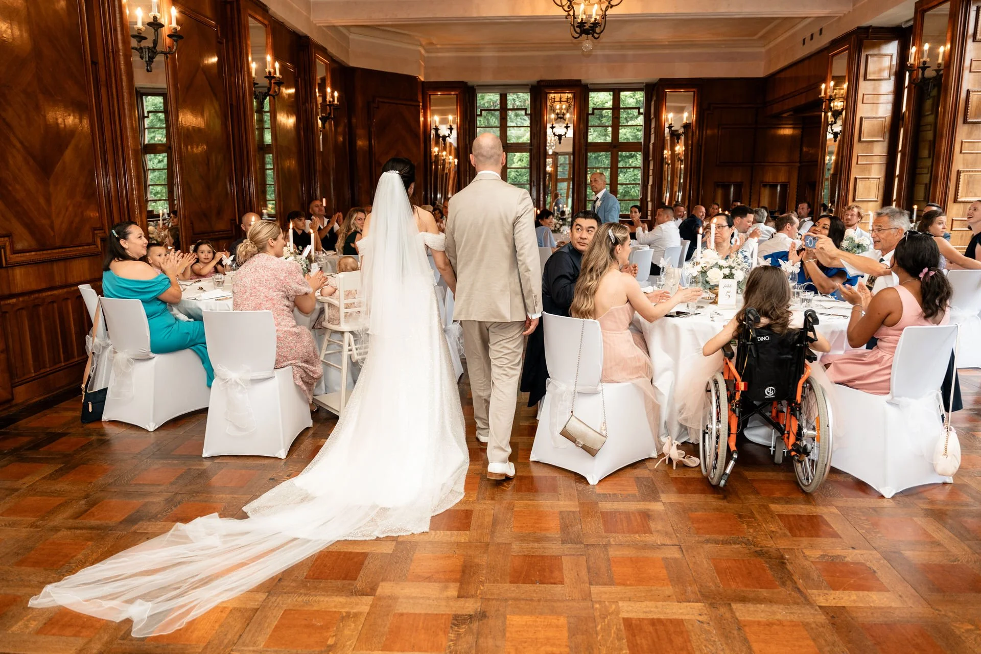 Hochzeitsgesellschaft in einem festlich gedeckten Saal, im Mittelpunkt ein Brautpaar, das beim Empfang in einem eleganten Raum mit Holzvertäfelung und großen Fenstern sitzt, umgeben von Gästen, einige in Rollstühlen, andere applaudieren.