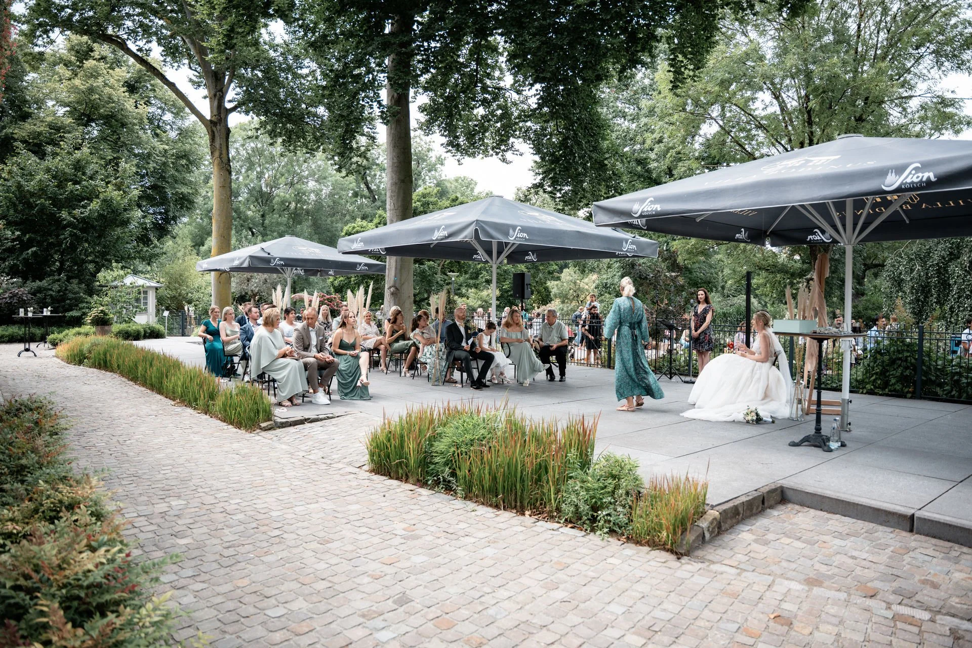Eine Hochzeitsfeier im Freien unter großen Sonnenschirmen mit Gästen, die auf Stühlen sitzen, während die Braut und ein Gast auf einer kleinen Bühne sprechen.