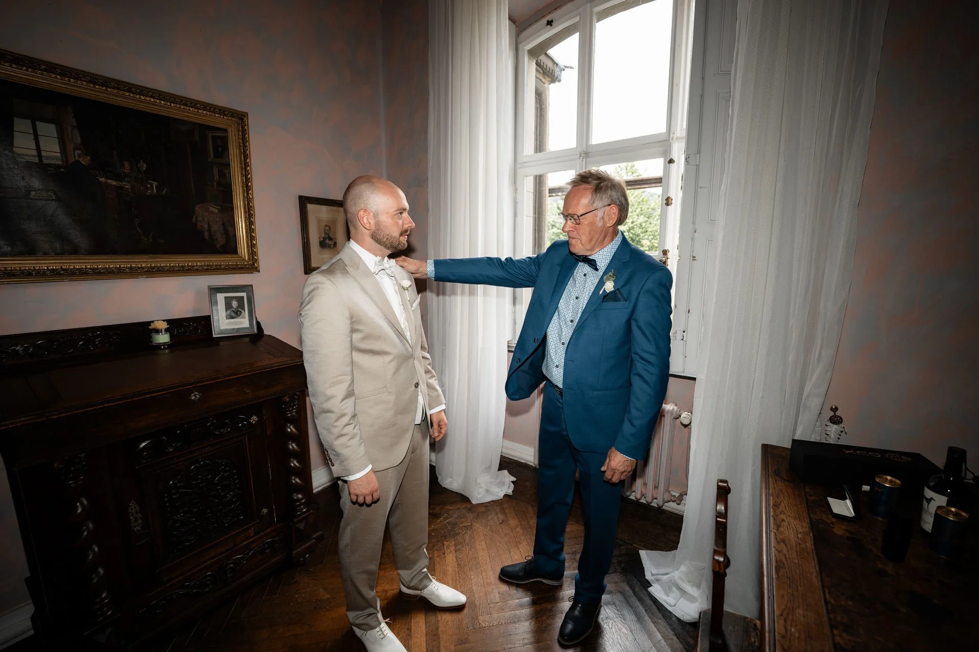 Zwei Männer im Anzug stehen sich gegenüber in einem Zimmer, der ältere Mann greift den jüngeren an die Schulter.