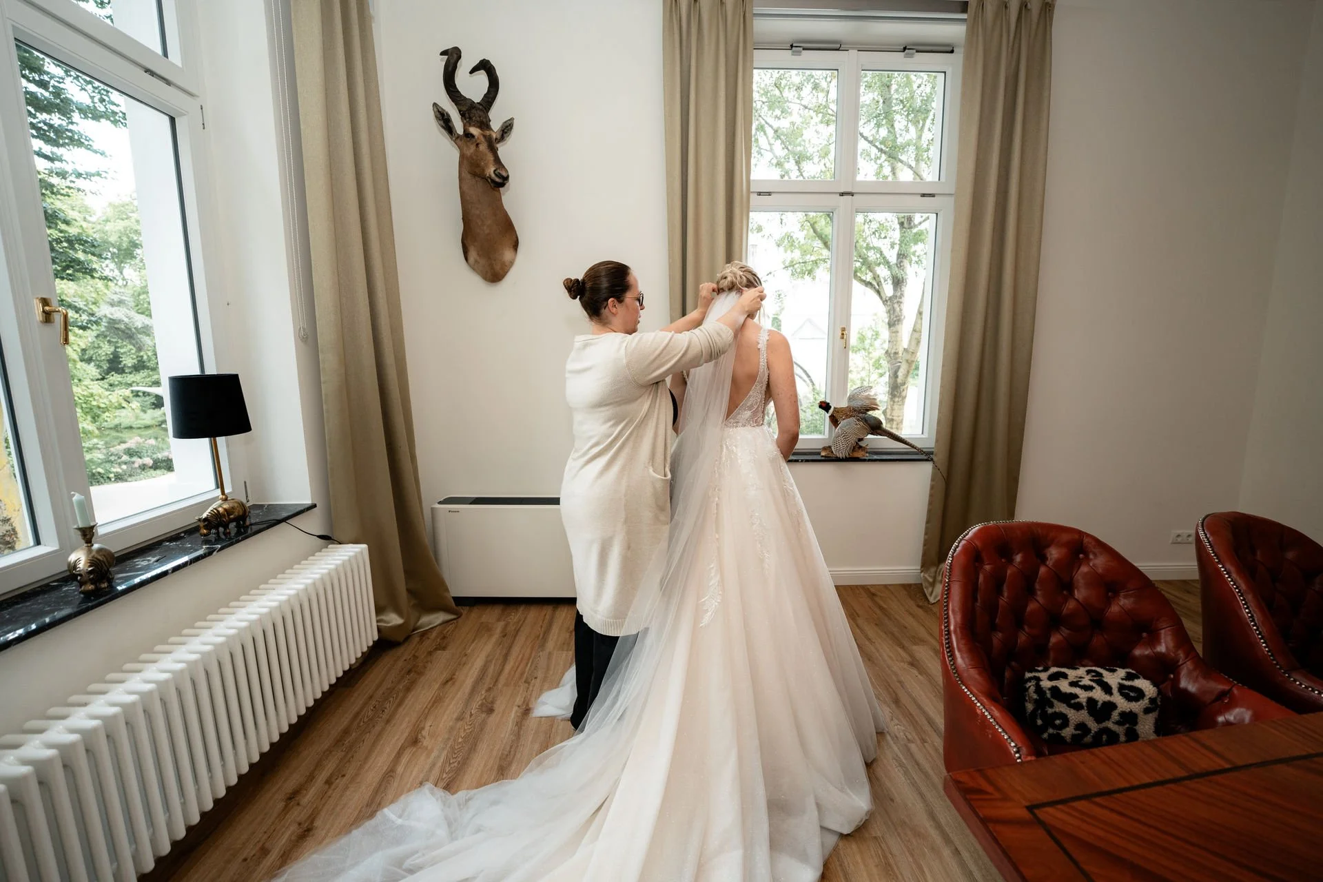 Eine Braut wird von einer Frau im weißen Kleid bei der Hochzeitsvorbereitung geholfen. Es ist ein helles Zimmer mit großen Fenstern und beige Vorhängen, Wanddekoration eines ziegenkopfs und gefedertem Vogel auf der Fensterbank.