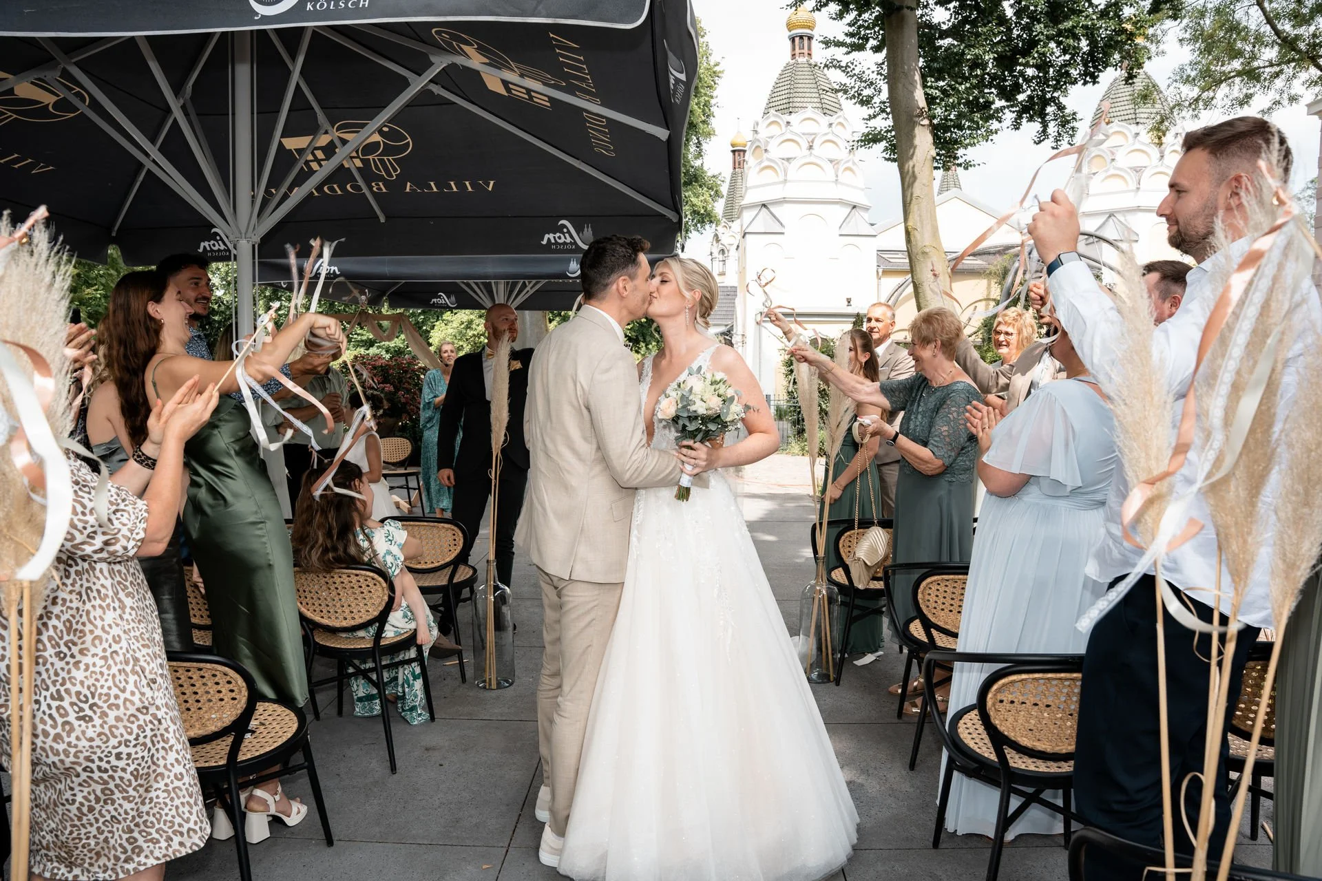 Ein frisch verheiratetes Paar küsst sich bei ihrer Hochzeit, umgeben von Freunden und Familie, die mit Luftschlangen feiern, im Freien vor einer Kirche.