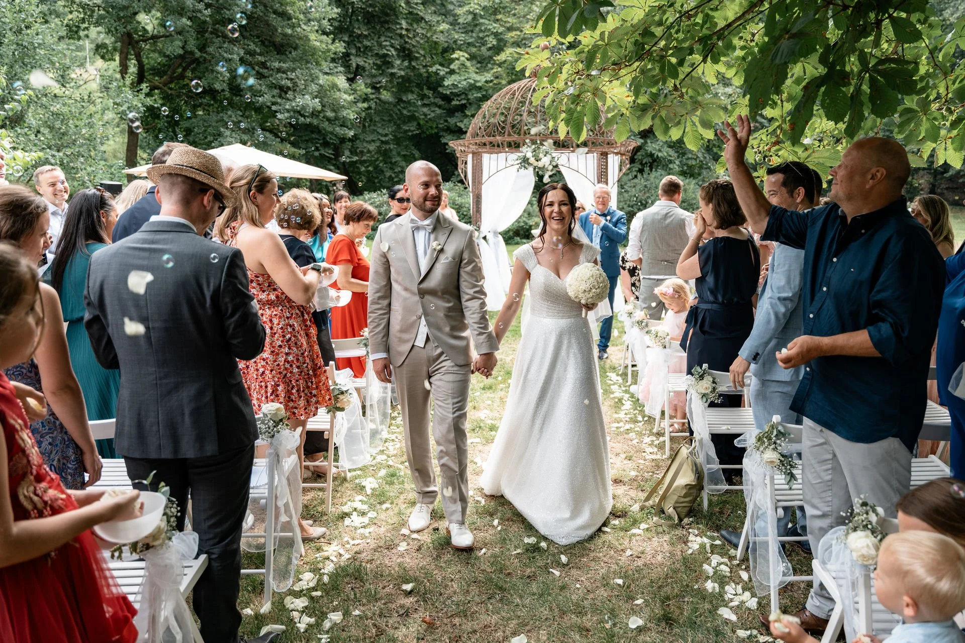 Ein glückliches Brautpaar geht den Gartenweg bei ihrer Hochzeit unter einer weiß geschmückten Laube entlang, umgeben von lachenden Gästen, wobei Blumenblätter in der Luft schweben.