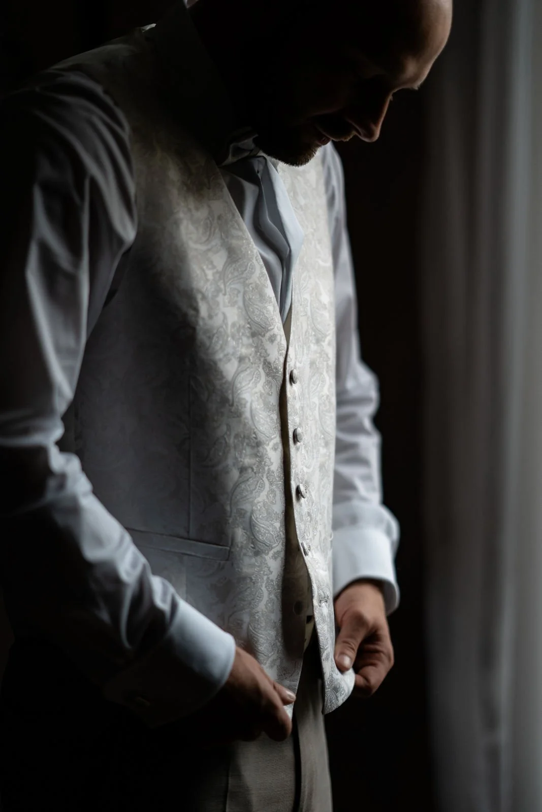 Mann adjusting seine Hochzeitsweste vor einem Fenster im Dunkeln.