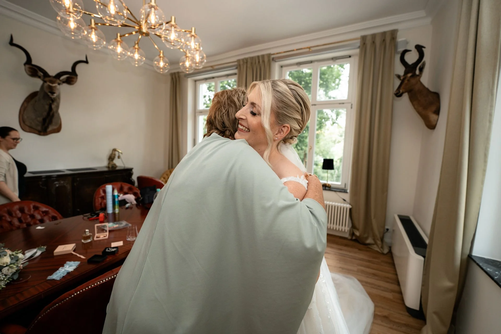 Braut umarmt eine Person in einem hellgrünen Kleid im eleganten Raum mit Kronleuchter und Wildtierköpfen an der Wand, während eine weitere Person im Hintergrund bei den Vorbereitungen hilft.