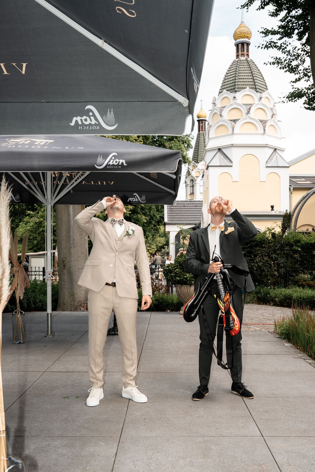 Zwei Männer in formeller Kleidung trinken aus kleinen Flugzeugen. Der Mann auf der linken trägt einen beigen Anzug mit Blumenmuster-Hemd und Bowtie, während der Mann auf der rechten einen schwarzen Anzug mit cremefarbener Krawatte und einer Gitarre t