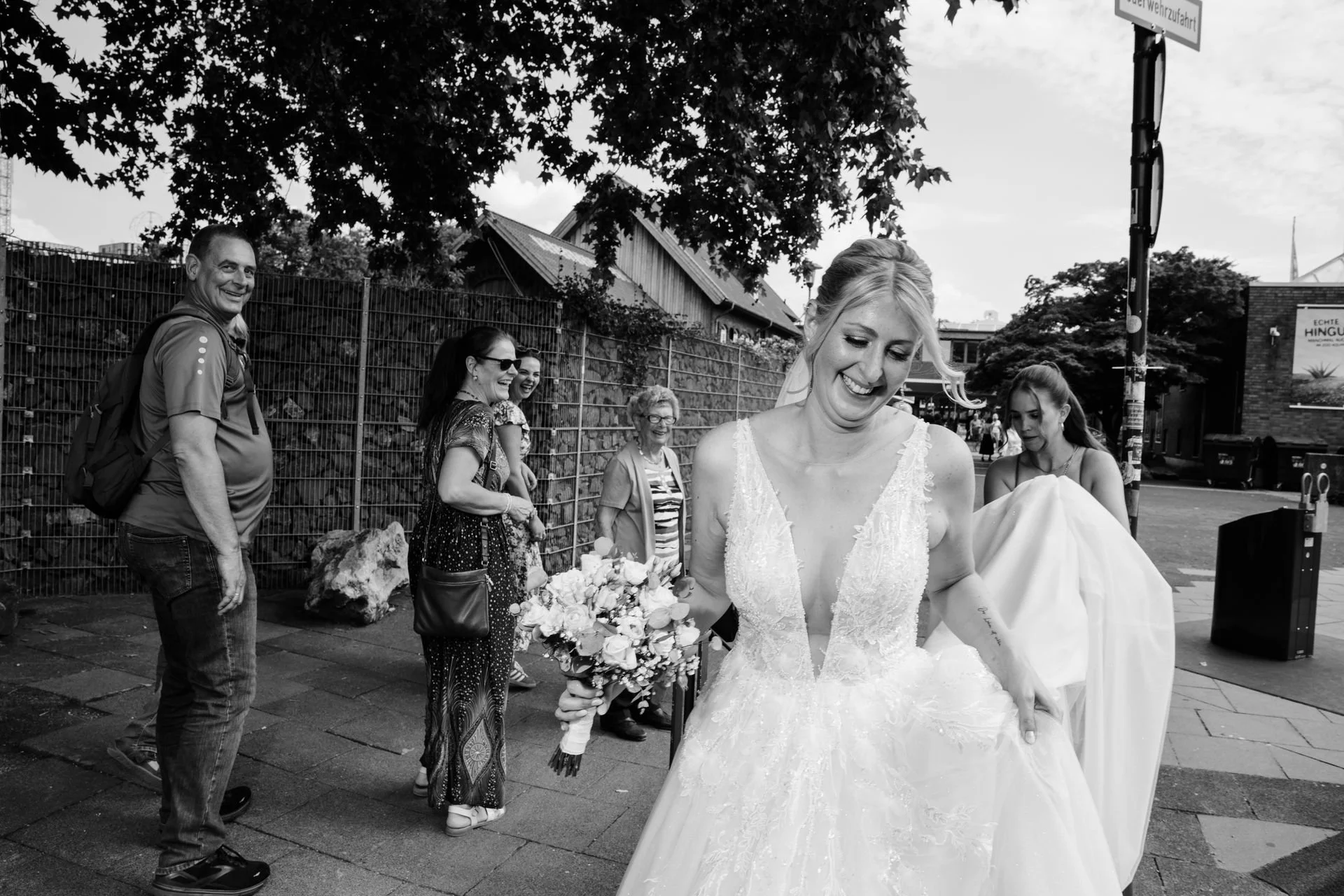 Eine lachende Braut in einem weißen Hochzeitskleid hält einen Blumenstrauß, umgeben von Freunden und Familie auf der Straße, im Hintergrund ein Zaun, Häuser und Bäume, die Szene ist in Schwarz-Weiß porträtiert.