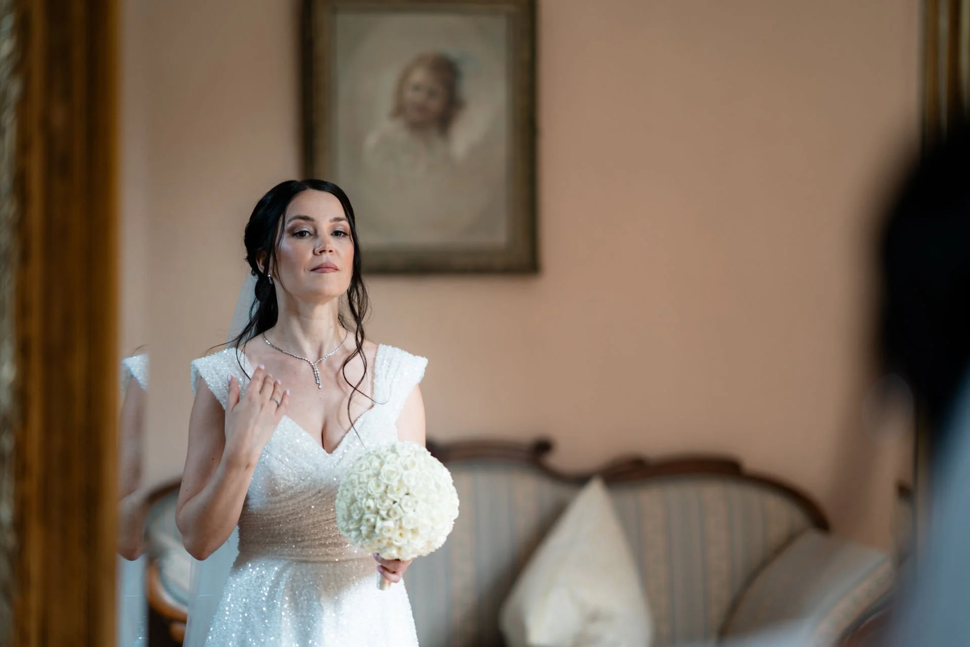 Eine Frau in einem weißen Hochzeitskleid hält einen Blumenstrauß und schaut lächelnd in einen Spiegel, während sie sich für ihre Hochzeit vorbereitet.