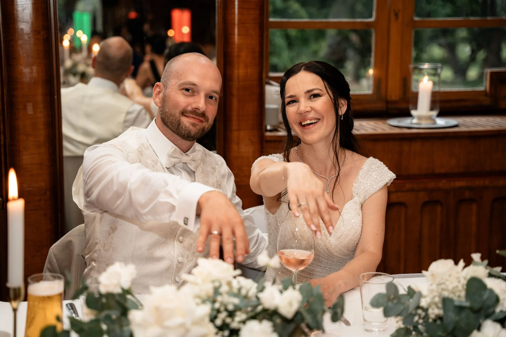 Ein glückliches Paar, wahrscheinlich Braut und Bräutigam, sitzt bei ihrer Hochzeitstafel, zeigt ihre Ringe und trinkt Sekt.