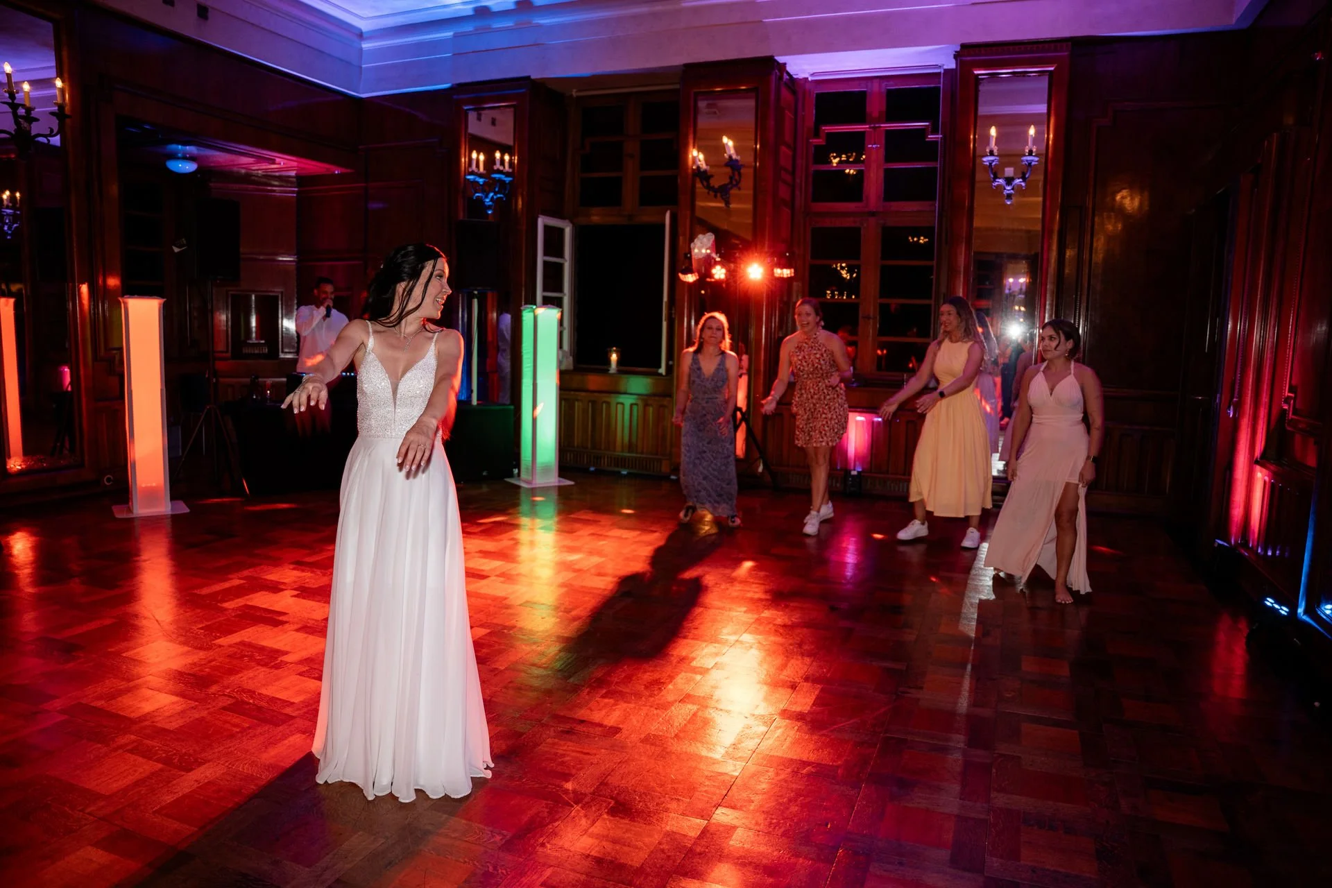 Frau in weißem Brautkleid tanzt in einem festlich beleuchteten Raum mit anderen Frauen im Hintergrund.