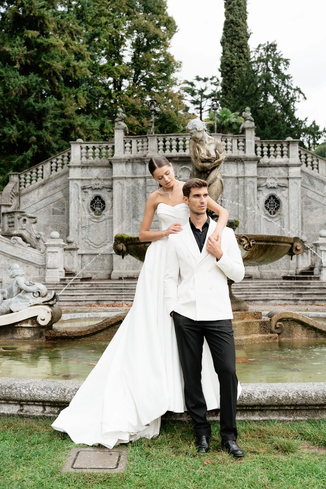 Ein Brautpaar posiert vor einem historischen Brunnen am Comer See, beide tragen elegante formelle Kleidung, die Frau in einem weißen Brautkleid und der Mann in einem weißen Anzug. Im Hintergrund sind grüne Bäume und eine Steintreppe sichtbar.
