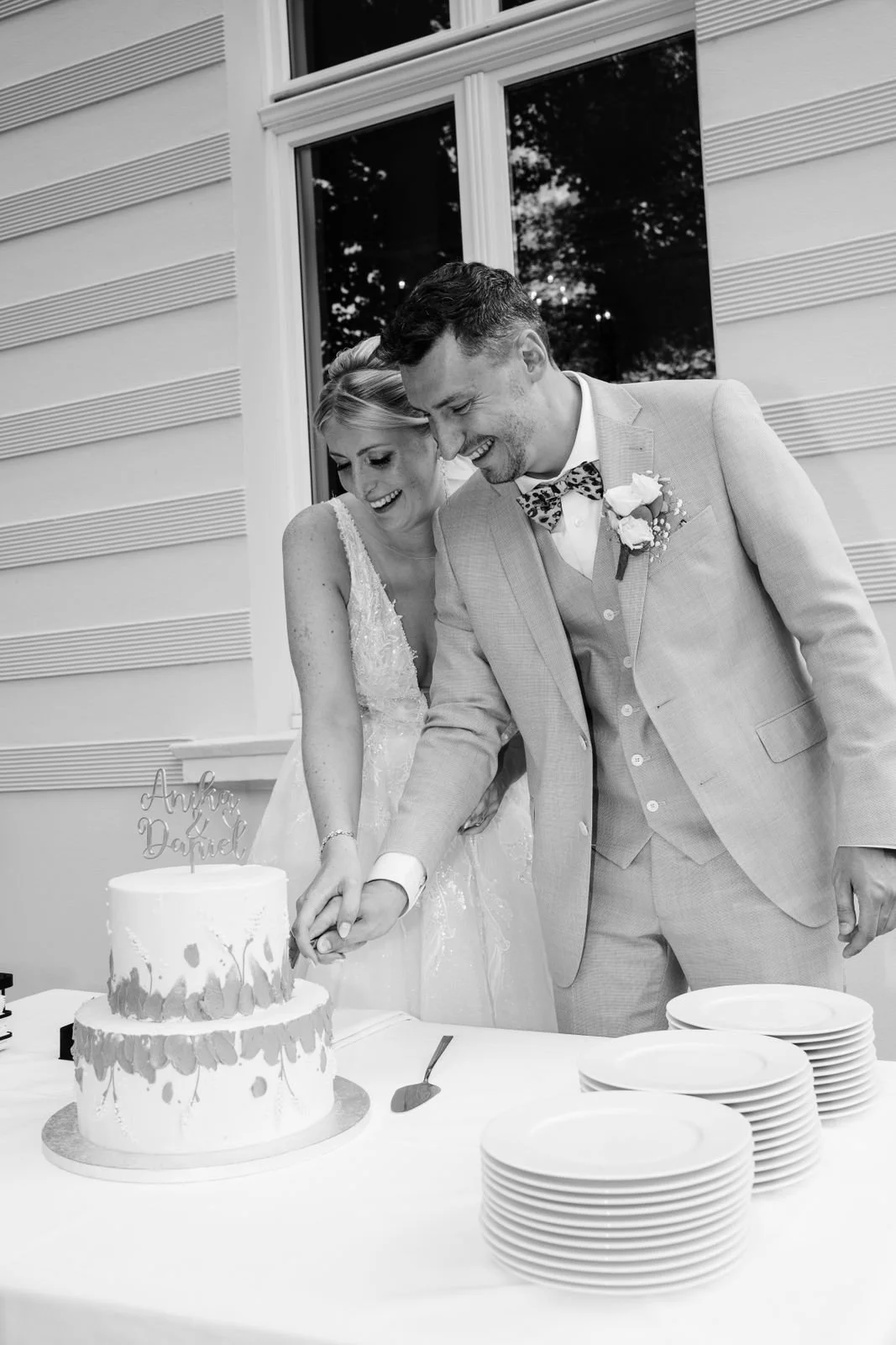 Braut und Bräutigam beim Anschneiden der Hochzeitstorte, in Schwarz-Weiß, vor einem Fenster, mehrere Teller auf dem Tisch.
