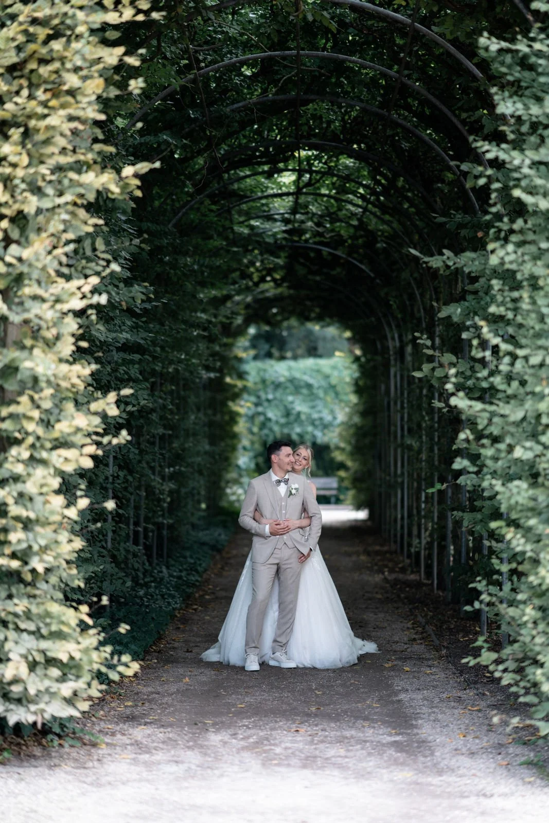 Ein Brautpaar steht in einem bewachsenen Tunnel aus Büschen und Bäumen, das in einem Park oder Garten liegt.