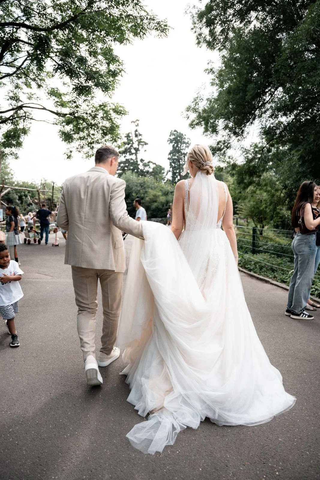 Ein Brautpaar geht Hand in Hand durch einen Park, umgeben von anderen Menschen und Bäumen, an einem Tag mit bewölktem Himmel.