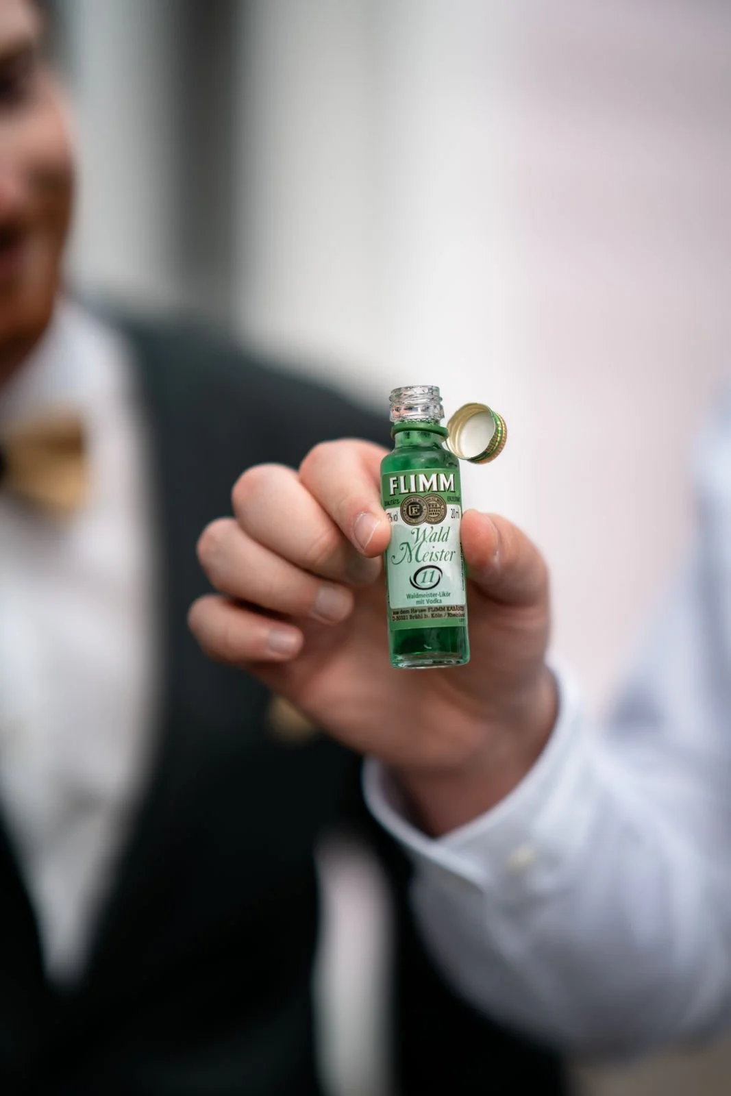 Nahaufnahme einer Hand, die eine kleine grüne Flasche mit transparentem Inhalt hält, mit einem geöffneten goldenen Verschluss. Im Hintergrund ist eine eingeroffene Person im Anzug sichtbar, aber unscharf.